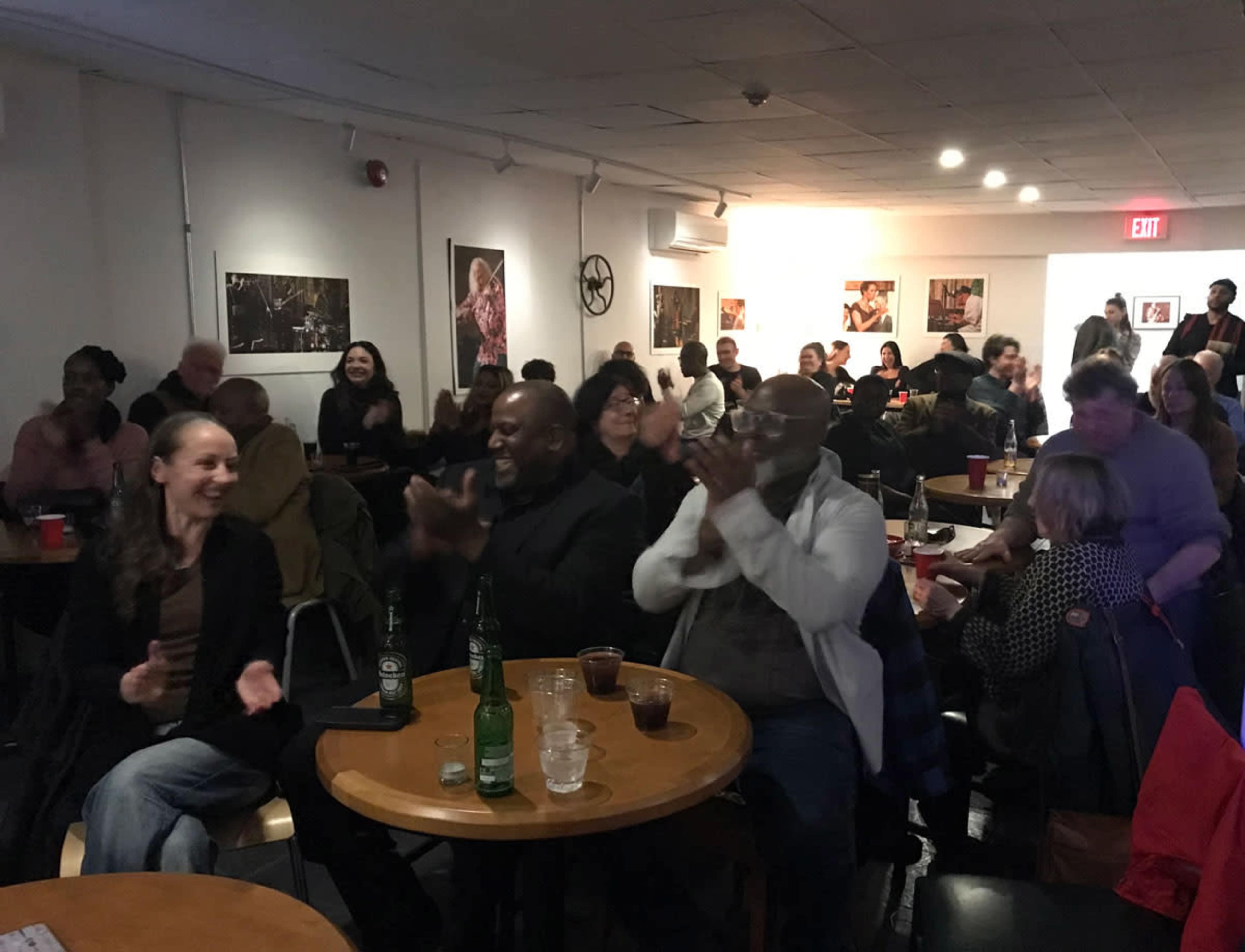A diverse crowd of people sits at tables in a warmly lit venue, clapping and smiling during a performance.
