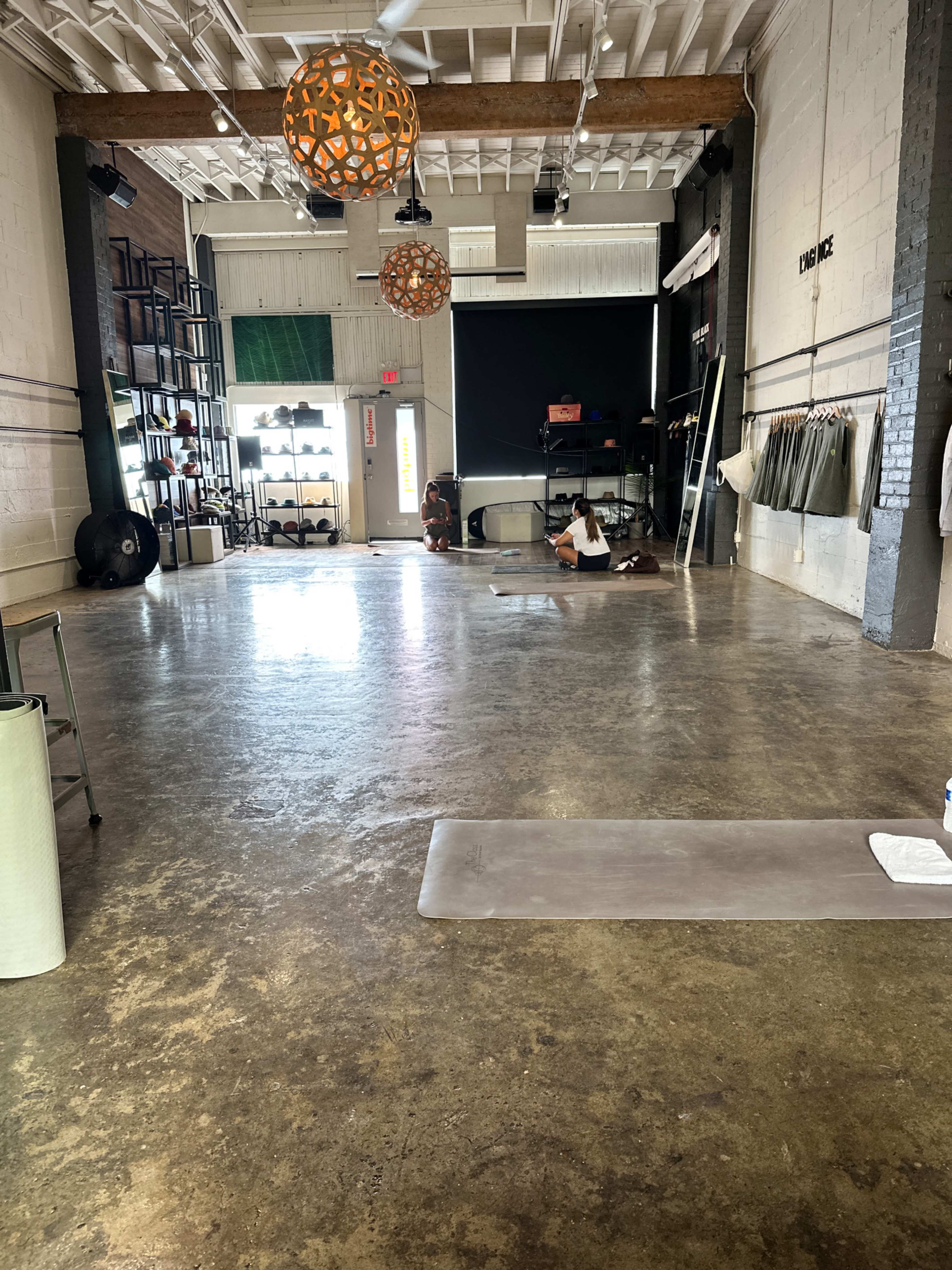 A spacious yoga studio with a polished concrete floor, large windows, and minimal decor, including two hanging light fixtures and yoga mats scattered across the area.