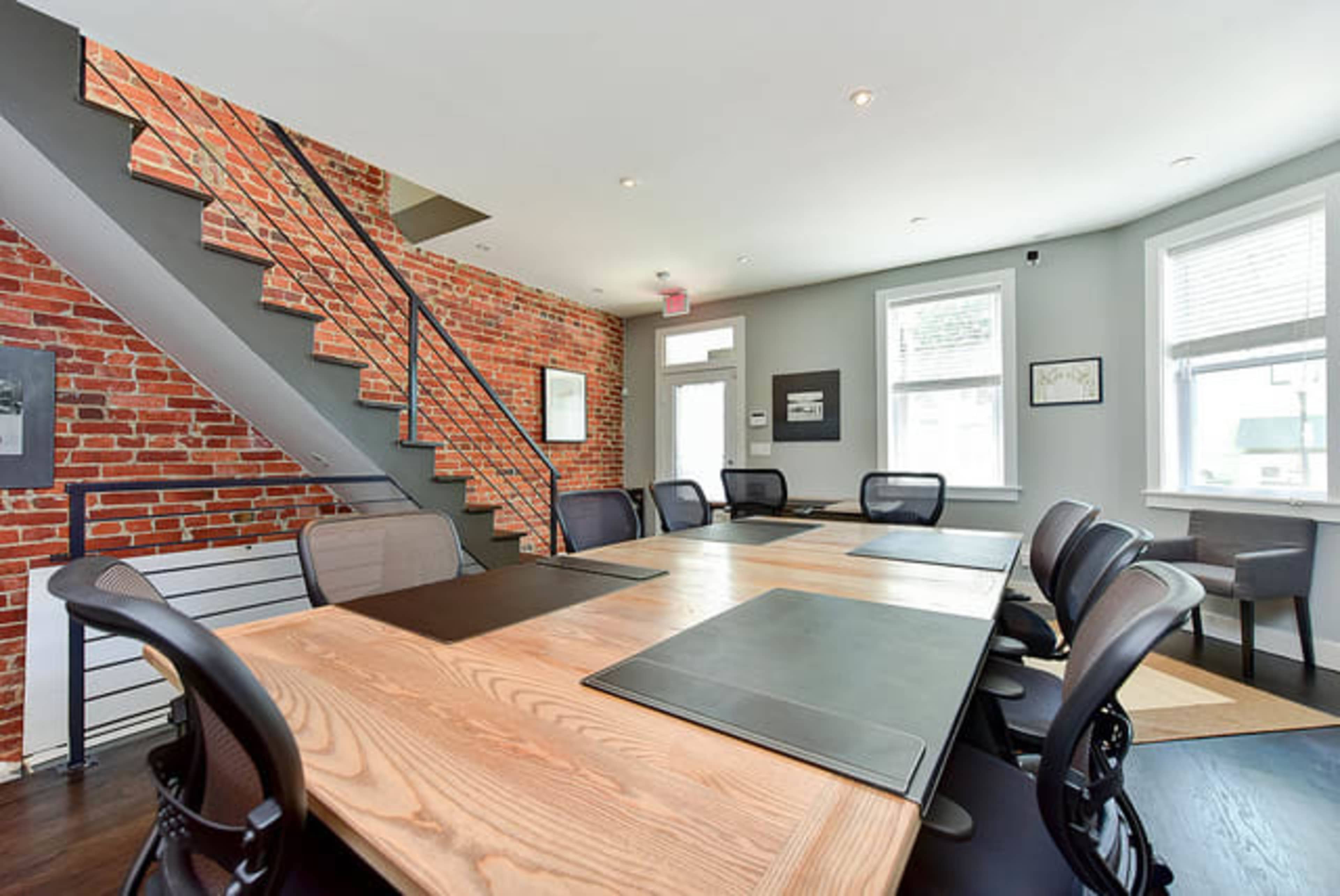 A modern conference room features a large wooden table surrounded by black office chairs, with exposed brick walls and a staircase in the background.