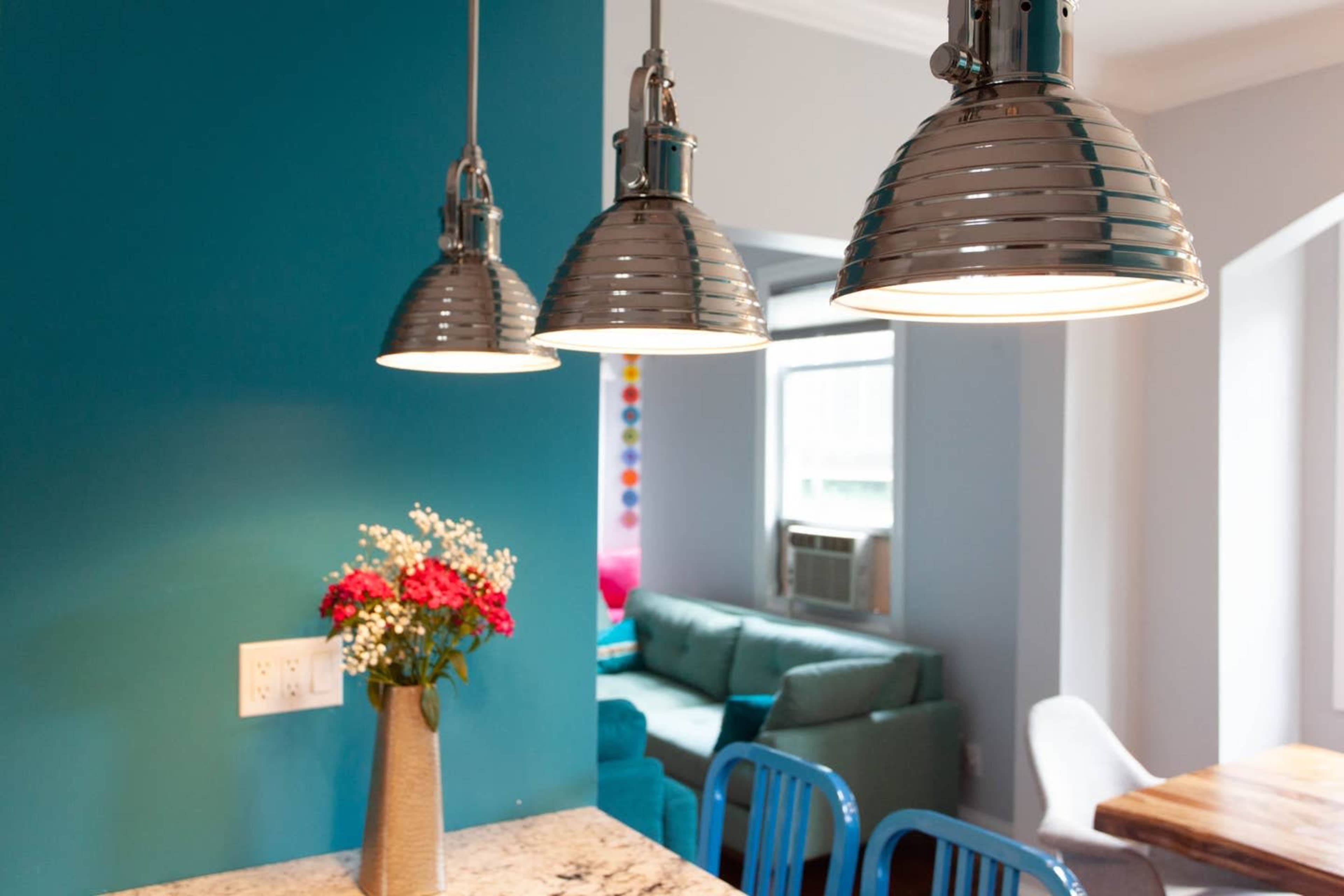 Three metal pendant lights hang above a kitchen counter, set against a teal wall, with a vase of flowers and a glimpse of a living area in the background.