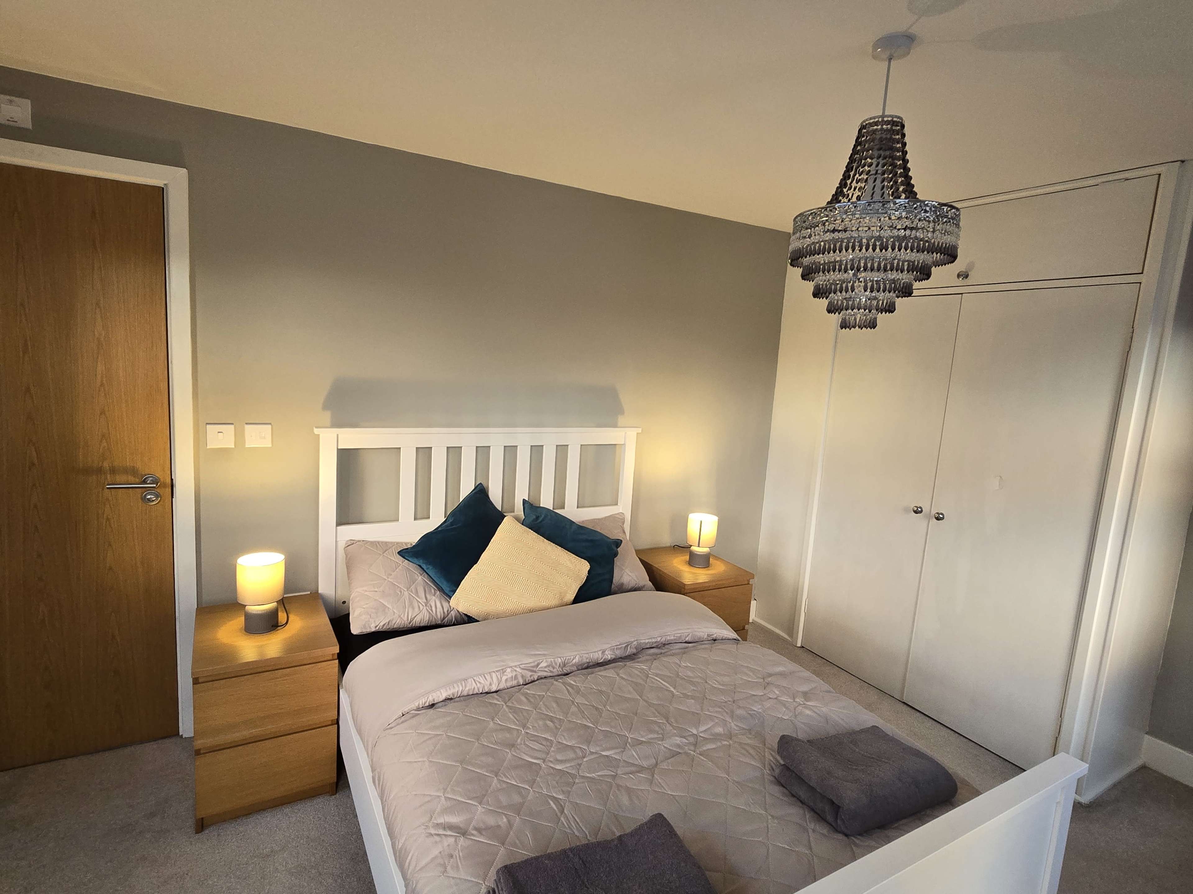 The image shows a well-decorated bedroom featuring a double bed with light bedding, two wooden nightstands, a chandelier, and a wardrobe.