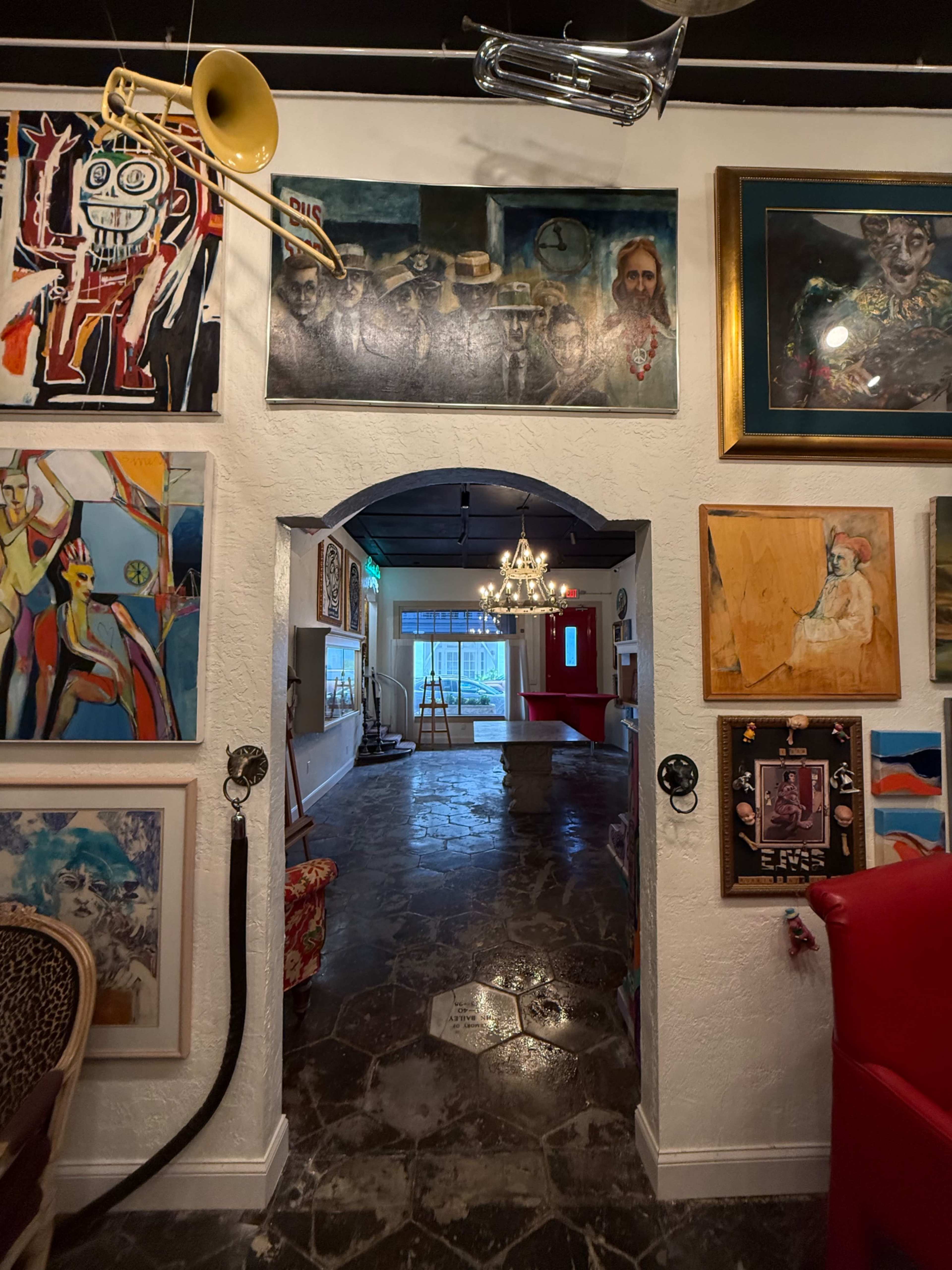 The image shows an indoor space with a variety of artworks displayed on white walls, leading through an archway towards a brightly lit area with a chandelier.