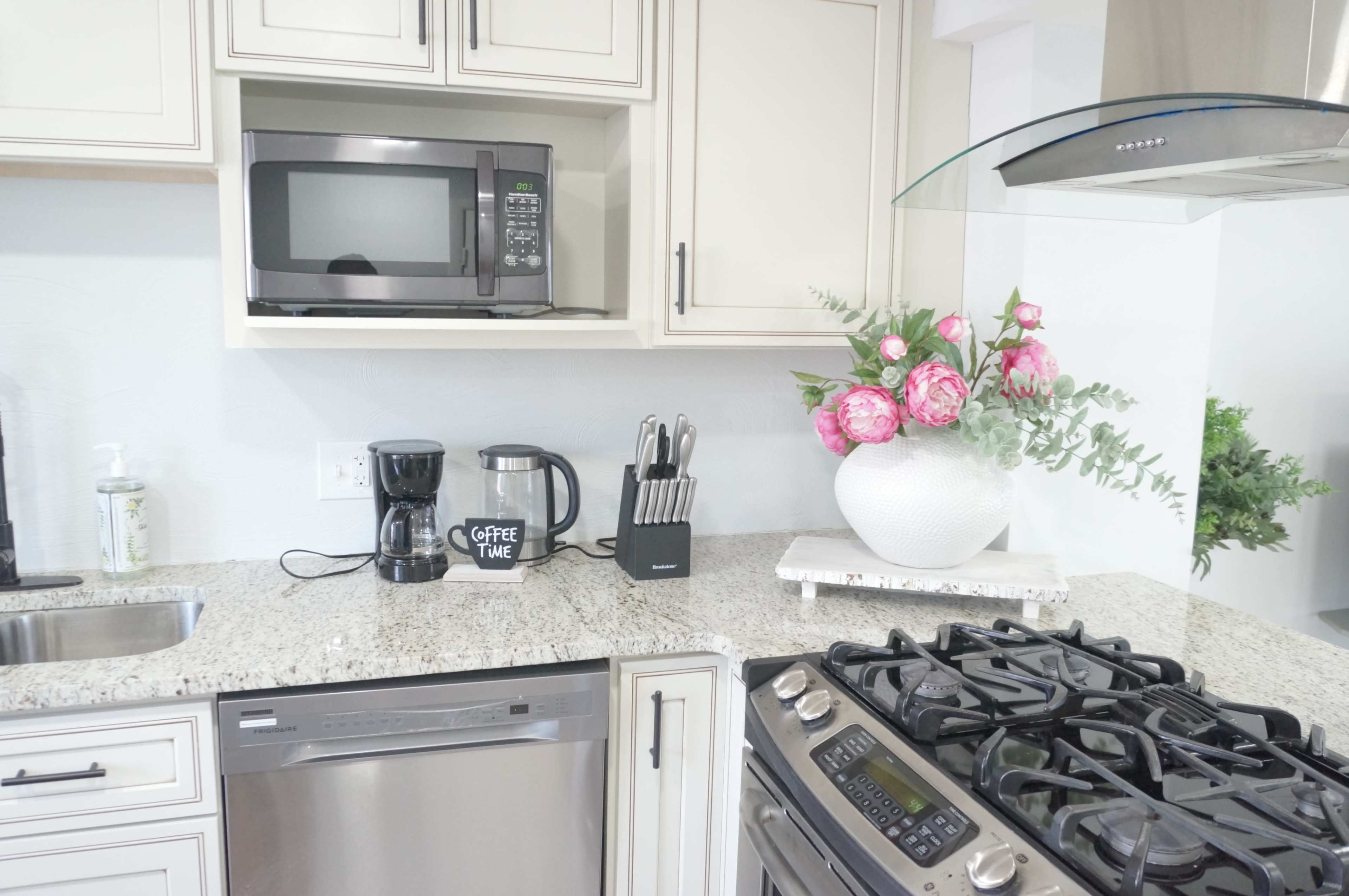 A modern kitchen features a granite countertop with a coffee maker, knife holder, and a microwave above, along with a floral arrangement and a gas stove.
