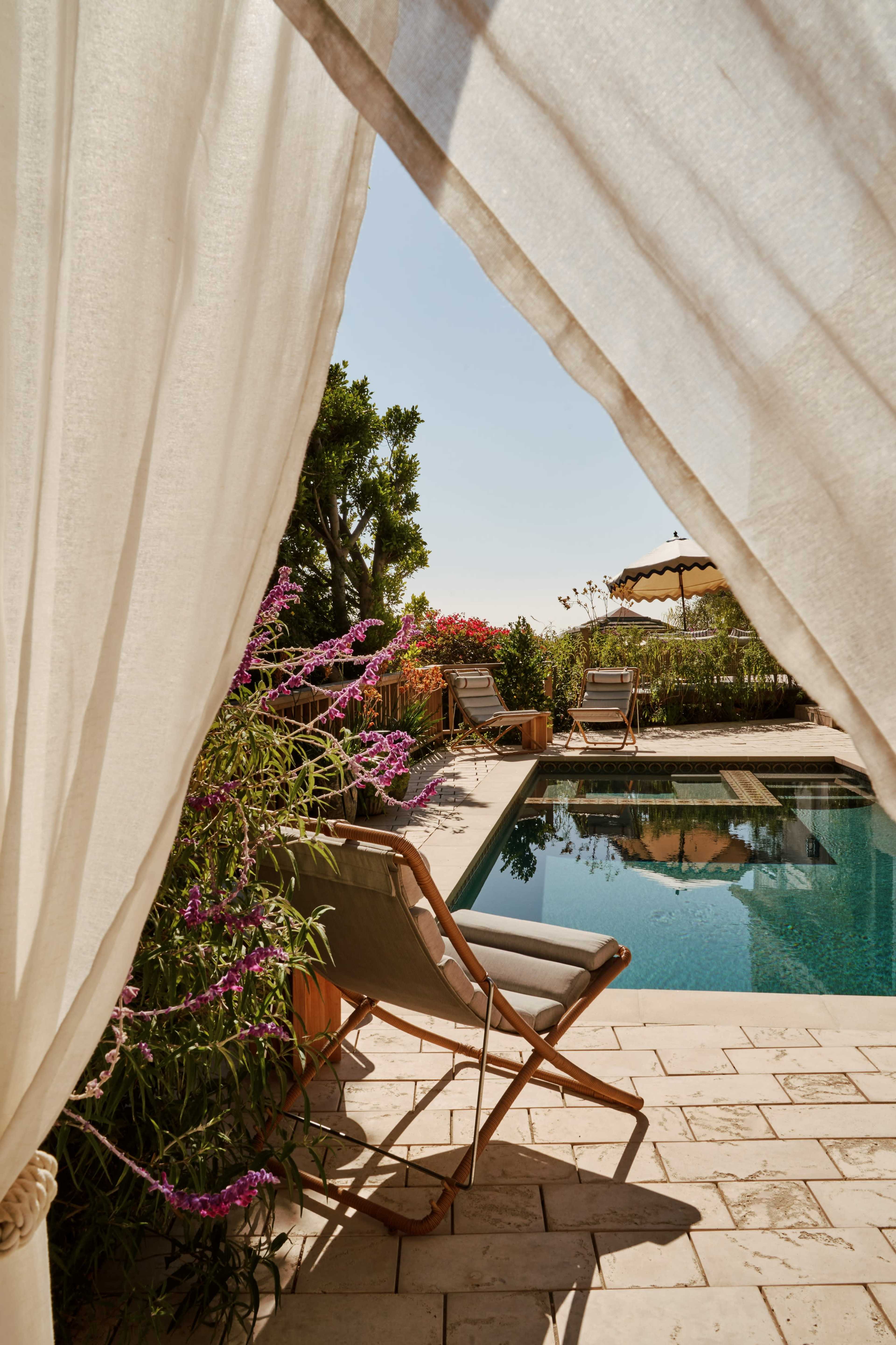 A view of a calm swimming pool surrounded by greenery and lounge chairs, framed by a flowing curtain.
