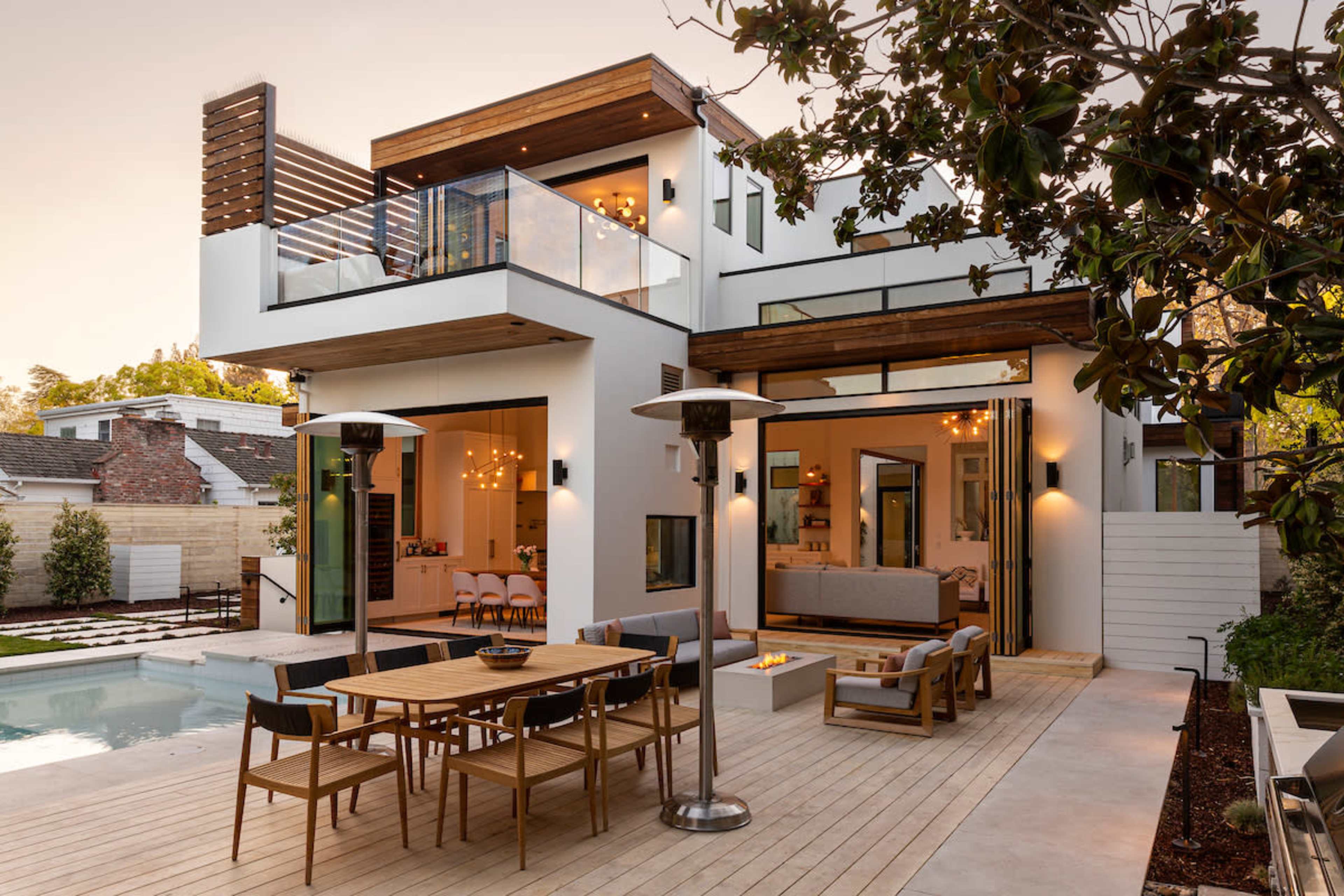 A modern two-story house features a spacious outdoor deck with a dining area, a pool, and large glass doors that connect to an interior living space.