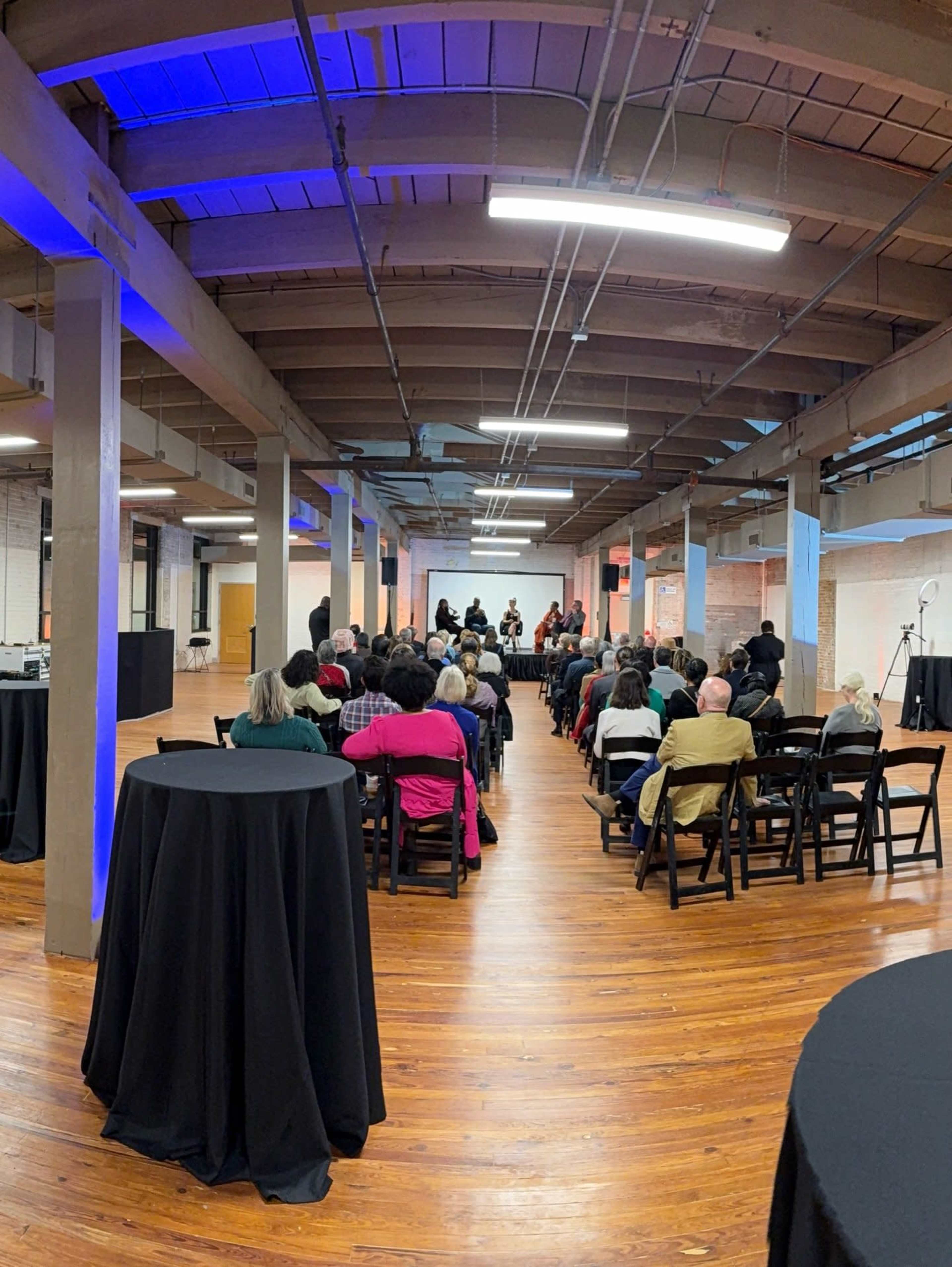 A group of people is seated in an industrial space with wooden floors, facing a panel on a stage under bright lights.