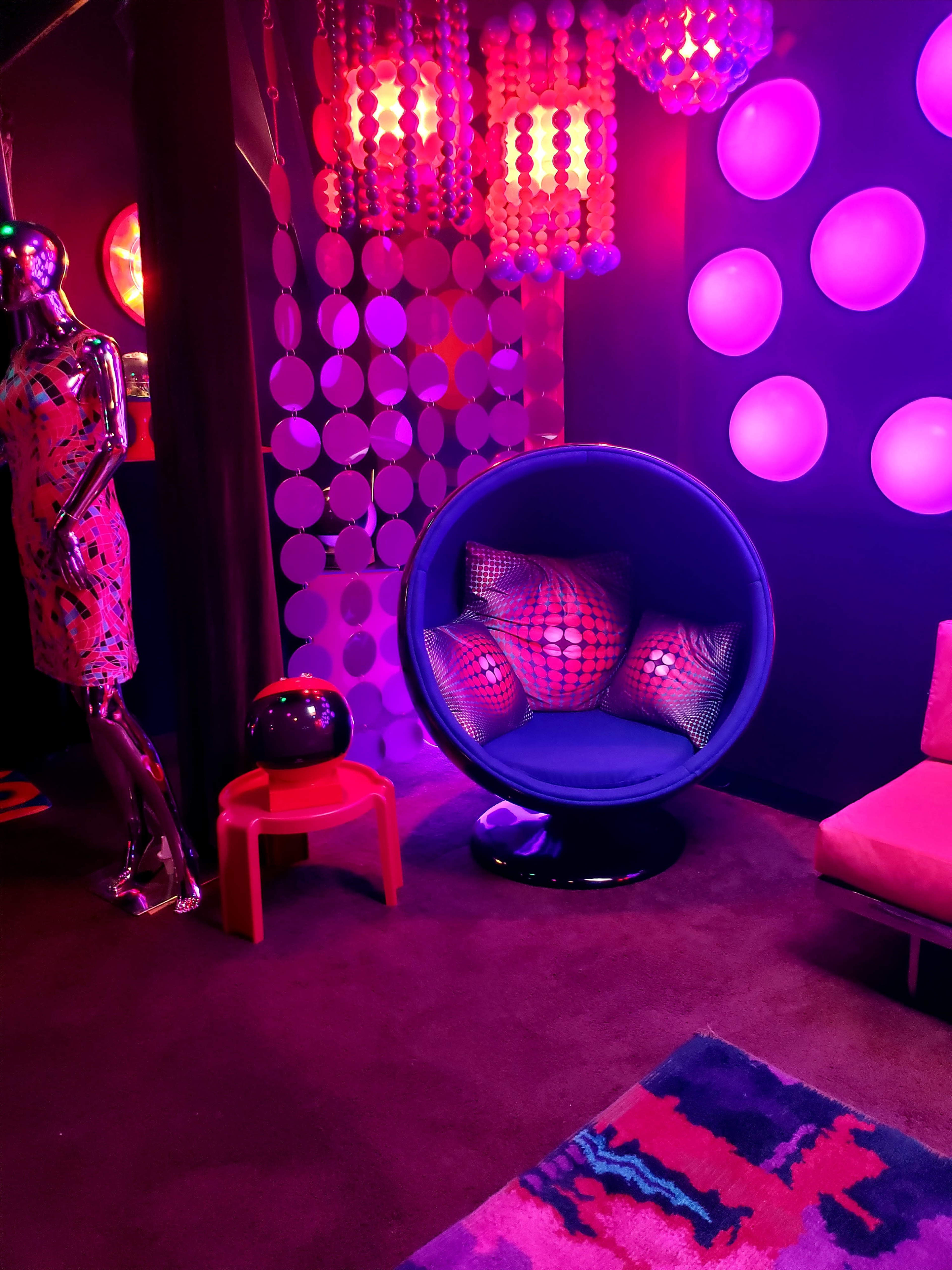 A vibrant retro-style lounge area featuring a blue pod chair, colorful decor, and a mannequin dressed in a patterned outfit.