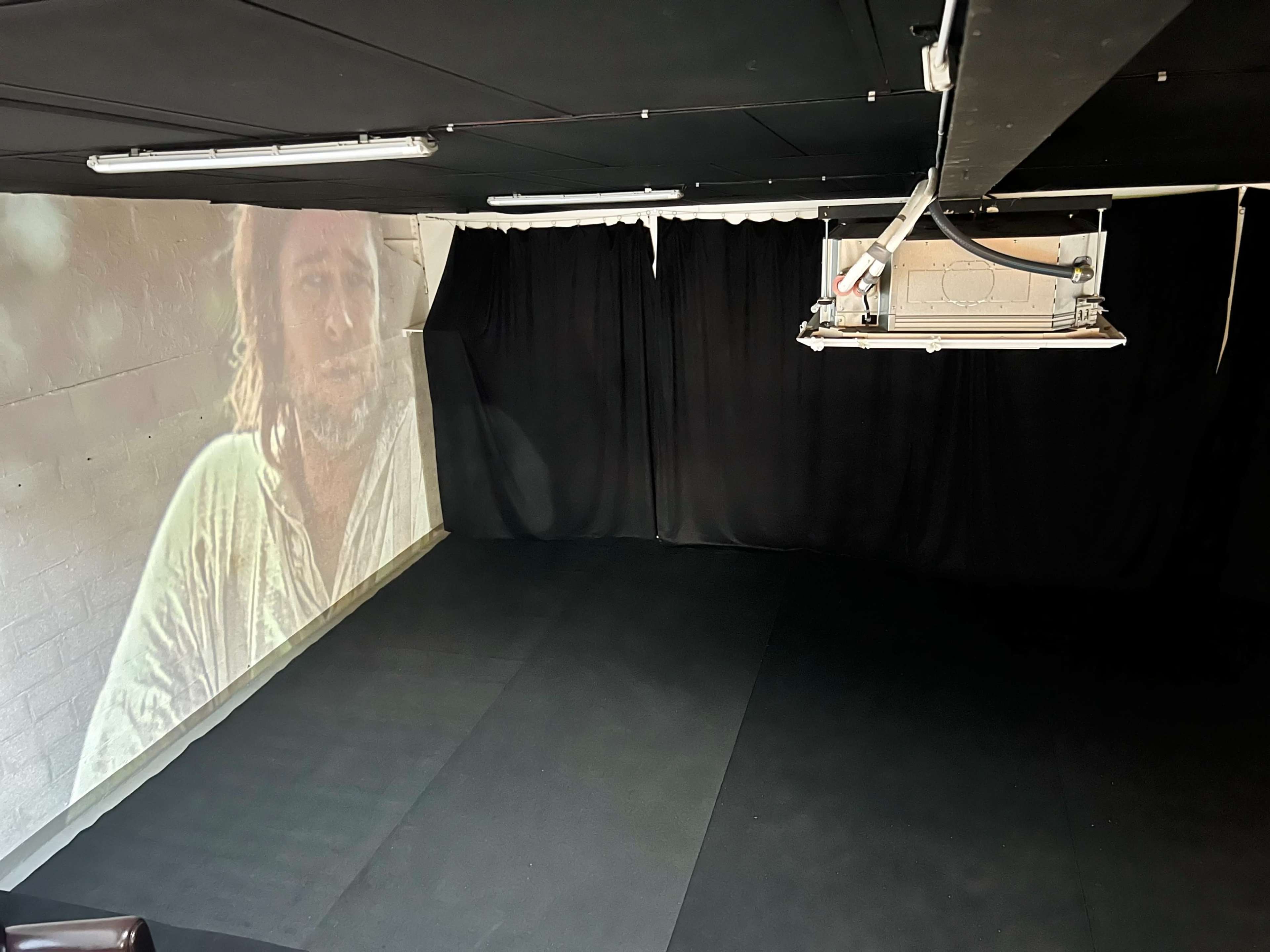 The image shows a dimly lit room with a large projection of a man’s face on one wall and black curtains covering other walls.