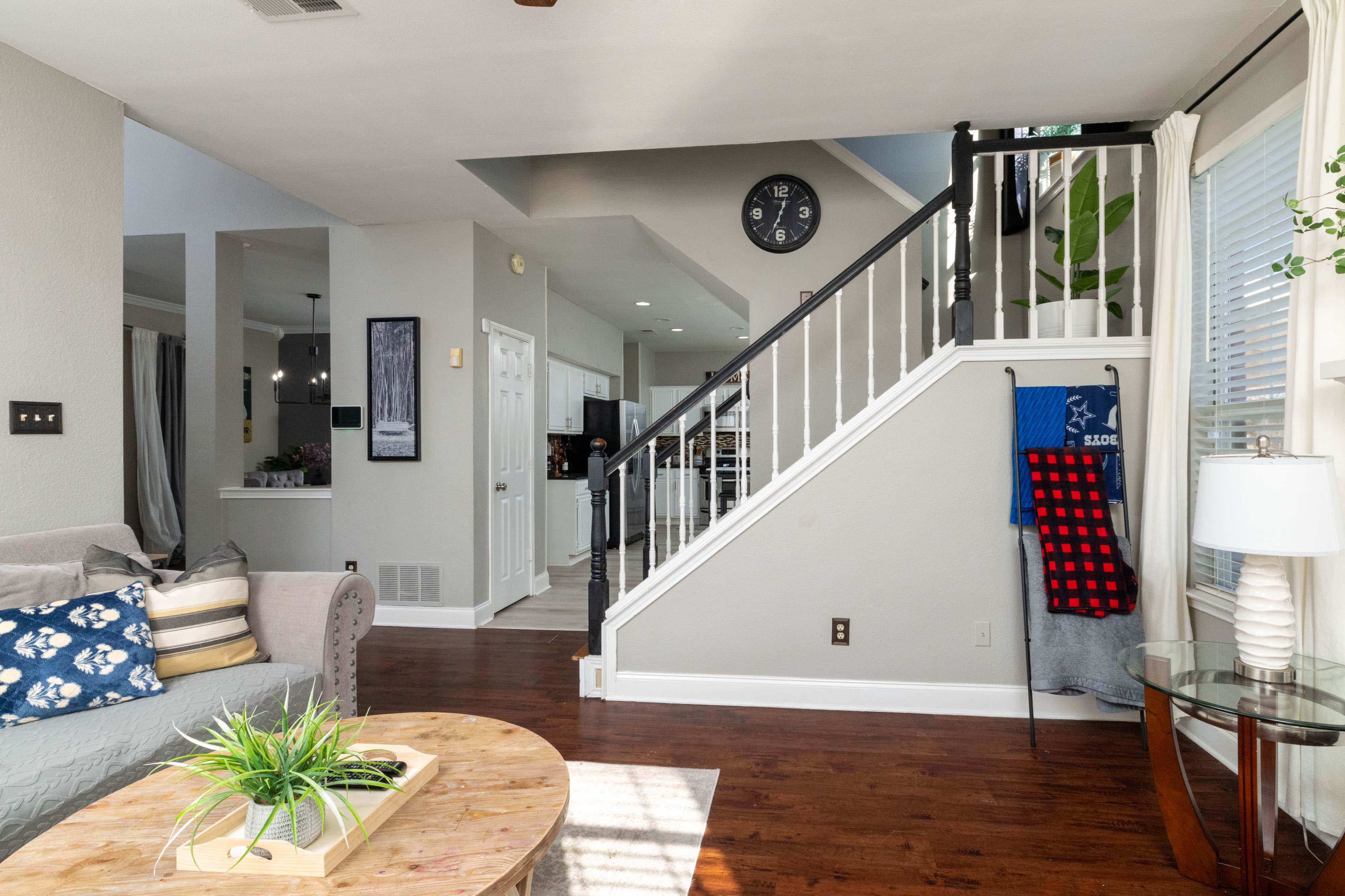 The image shows a modern living room with a staircase, a round wooden coffee table, and a wall clock above the stairs.