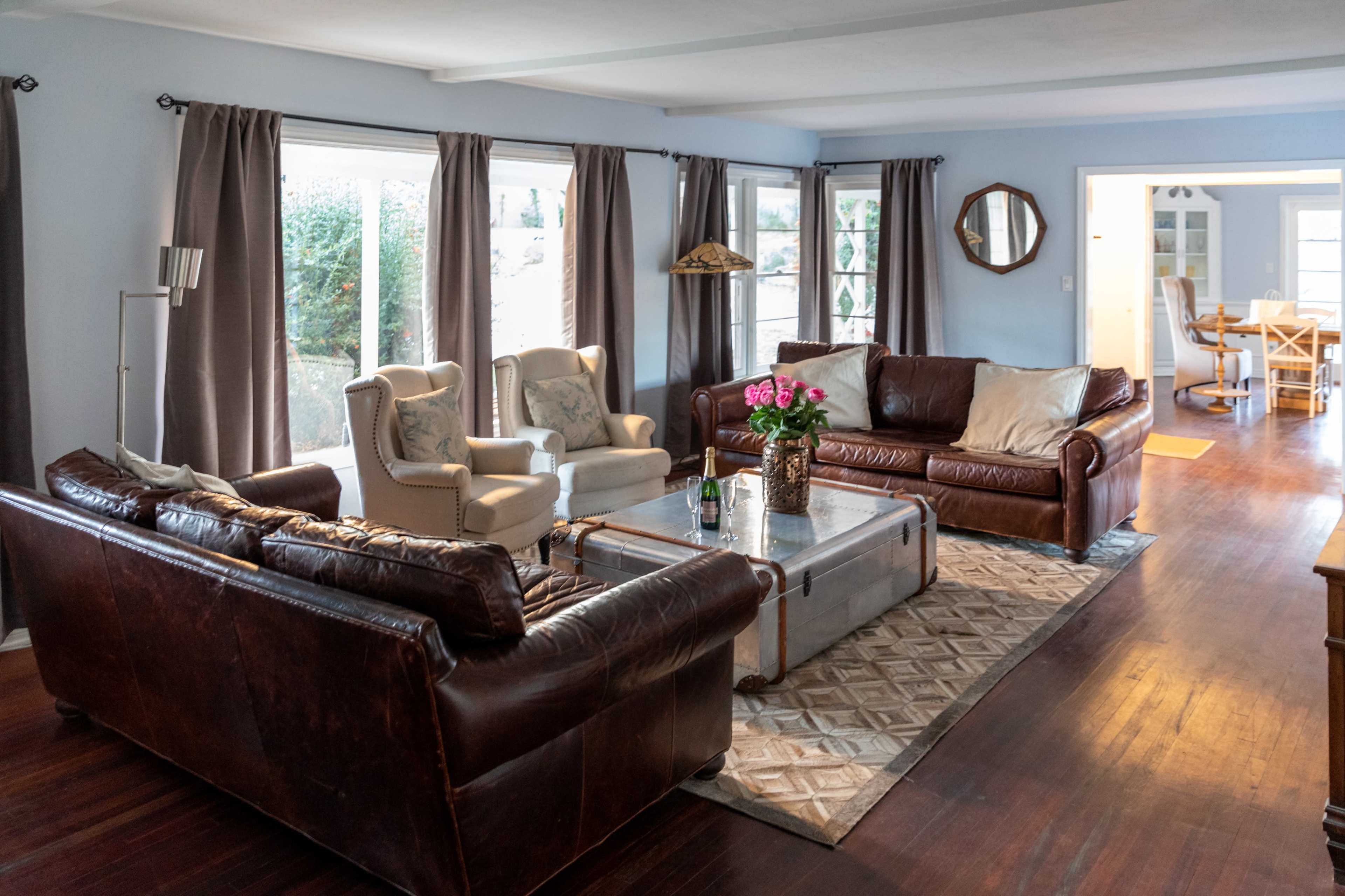 The living room features two leather sofas and two upholstered chairs arranged around a coffee table, with large windows allowing natural light to enter.