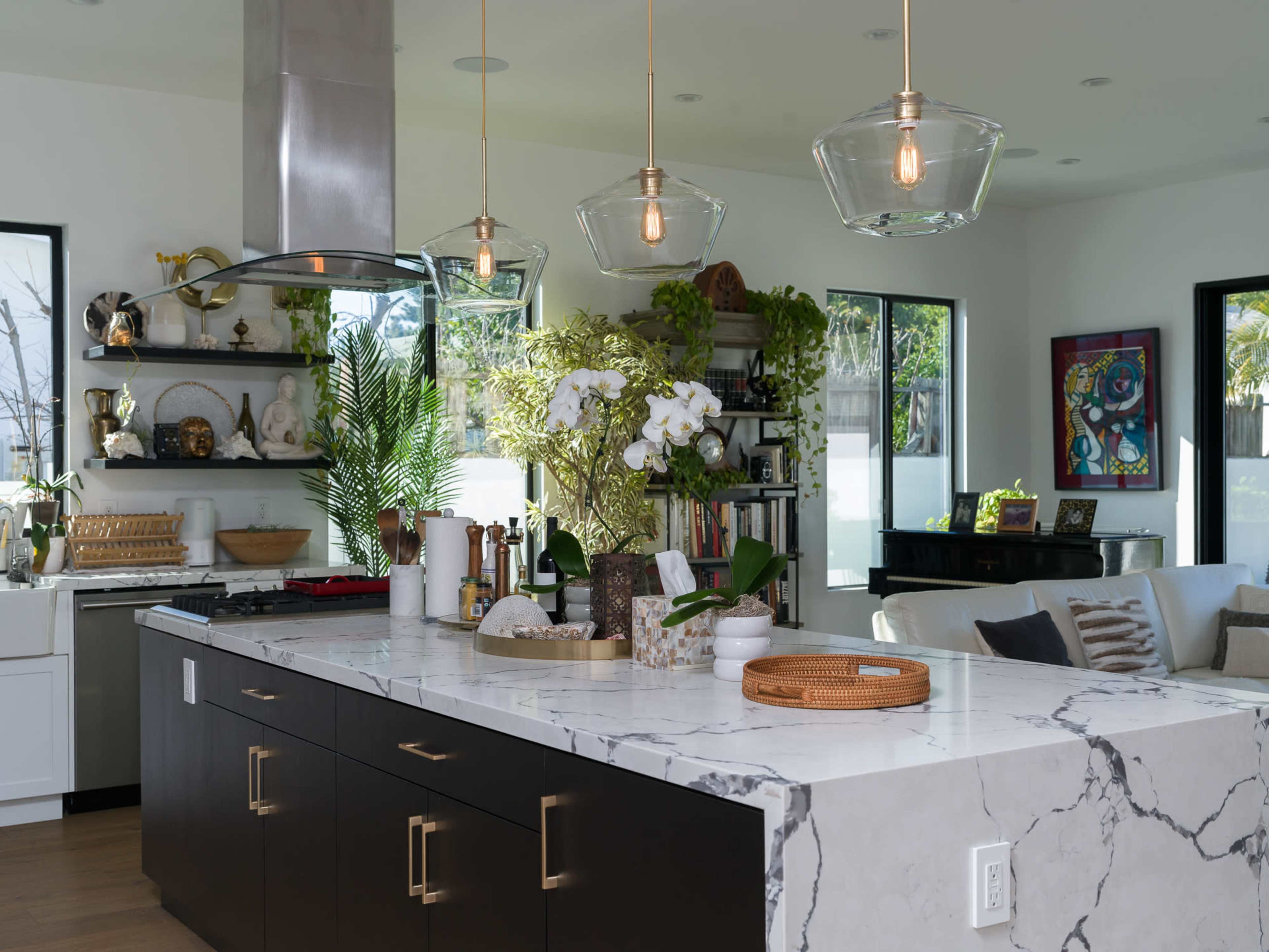 A modern kitchen features a large marble island with dark cabinets, plants, and decorative items, alongside open shelving and large windows.