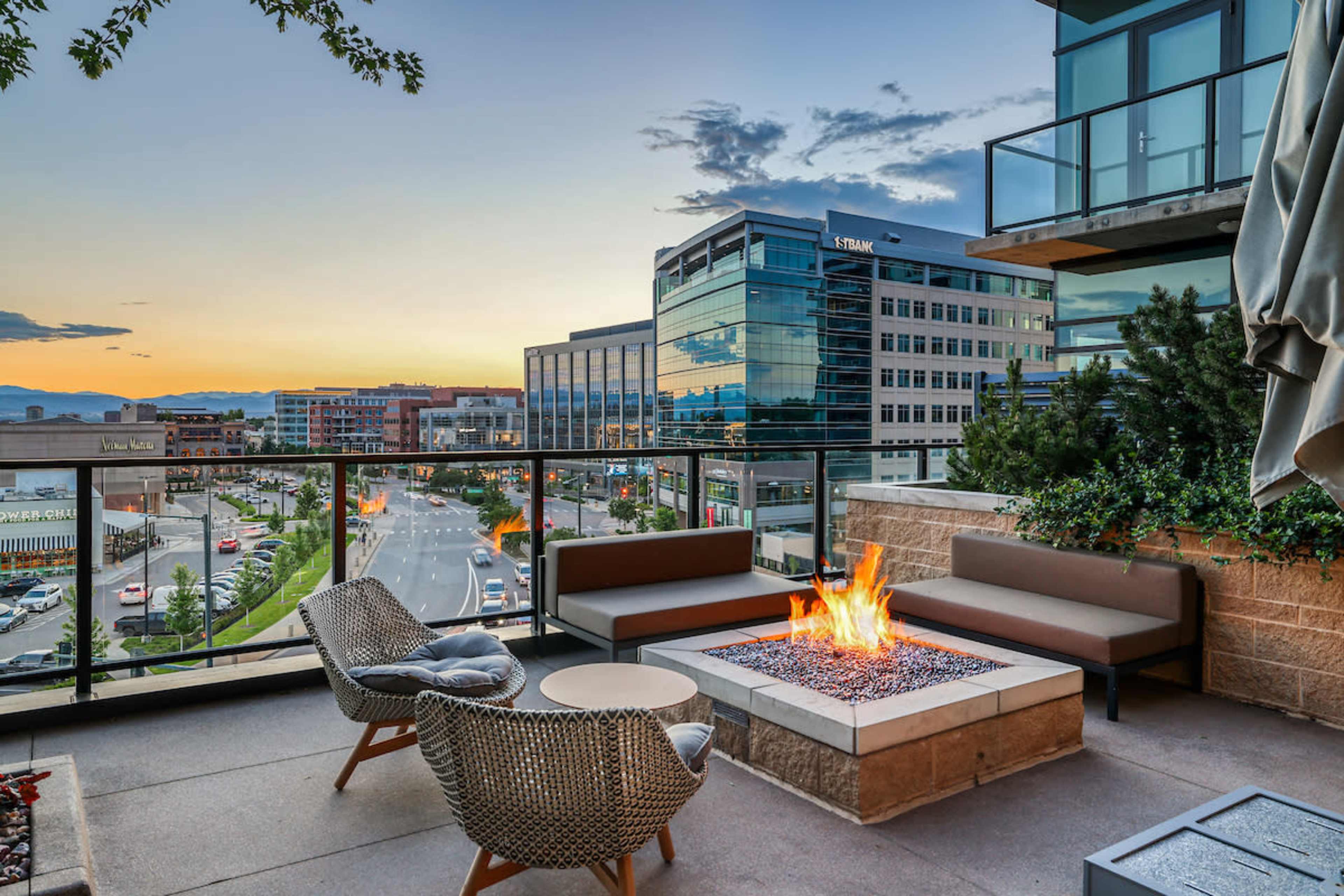 A rooftop patio features a modern fire pit surrounded by seating, overlooking a cityscape at sunset.