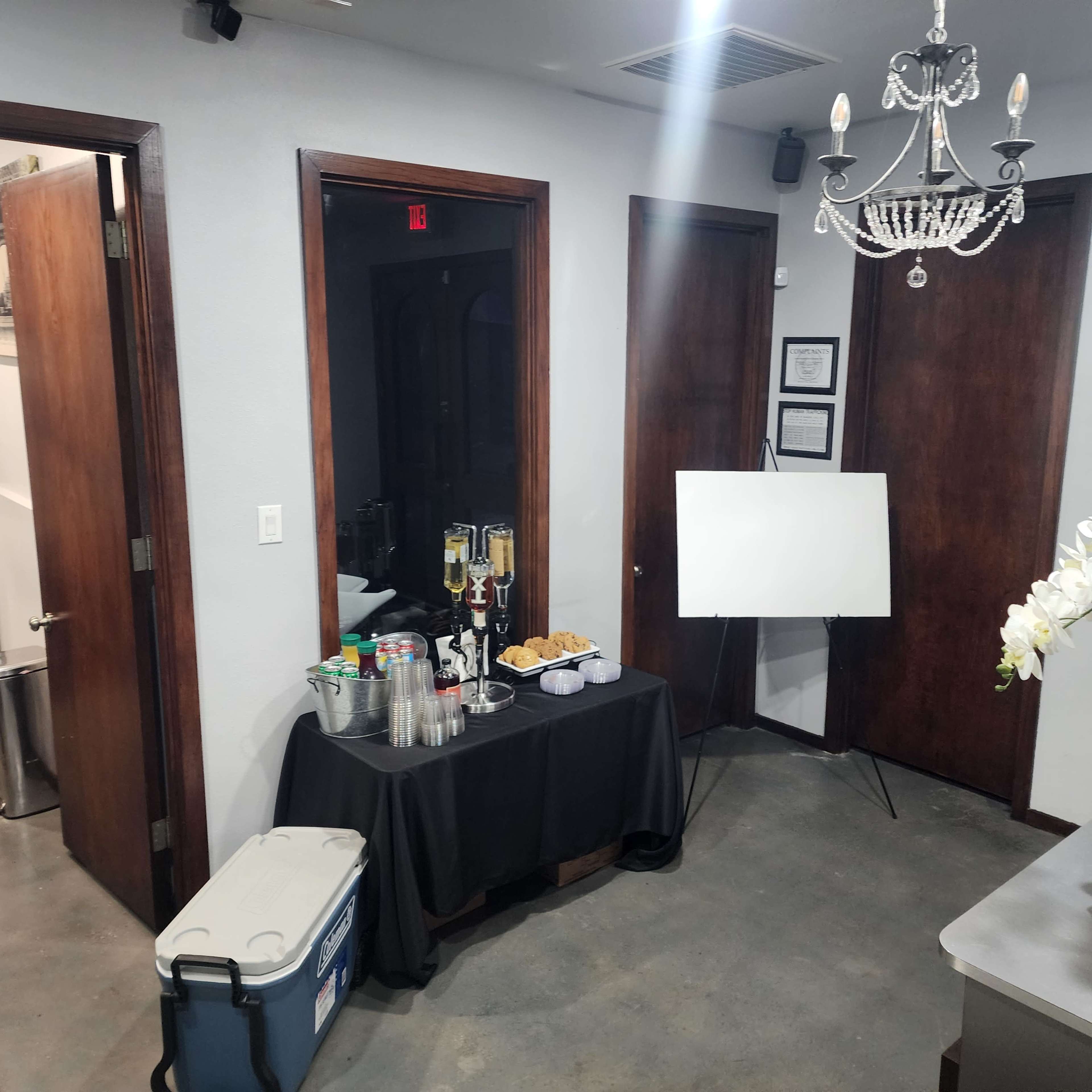 The image shows a small event space with two wooden doors, a table covered with a black cloth displaying drinks and snacks, and a blank easel on display.