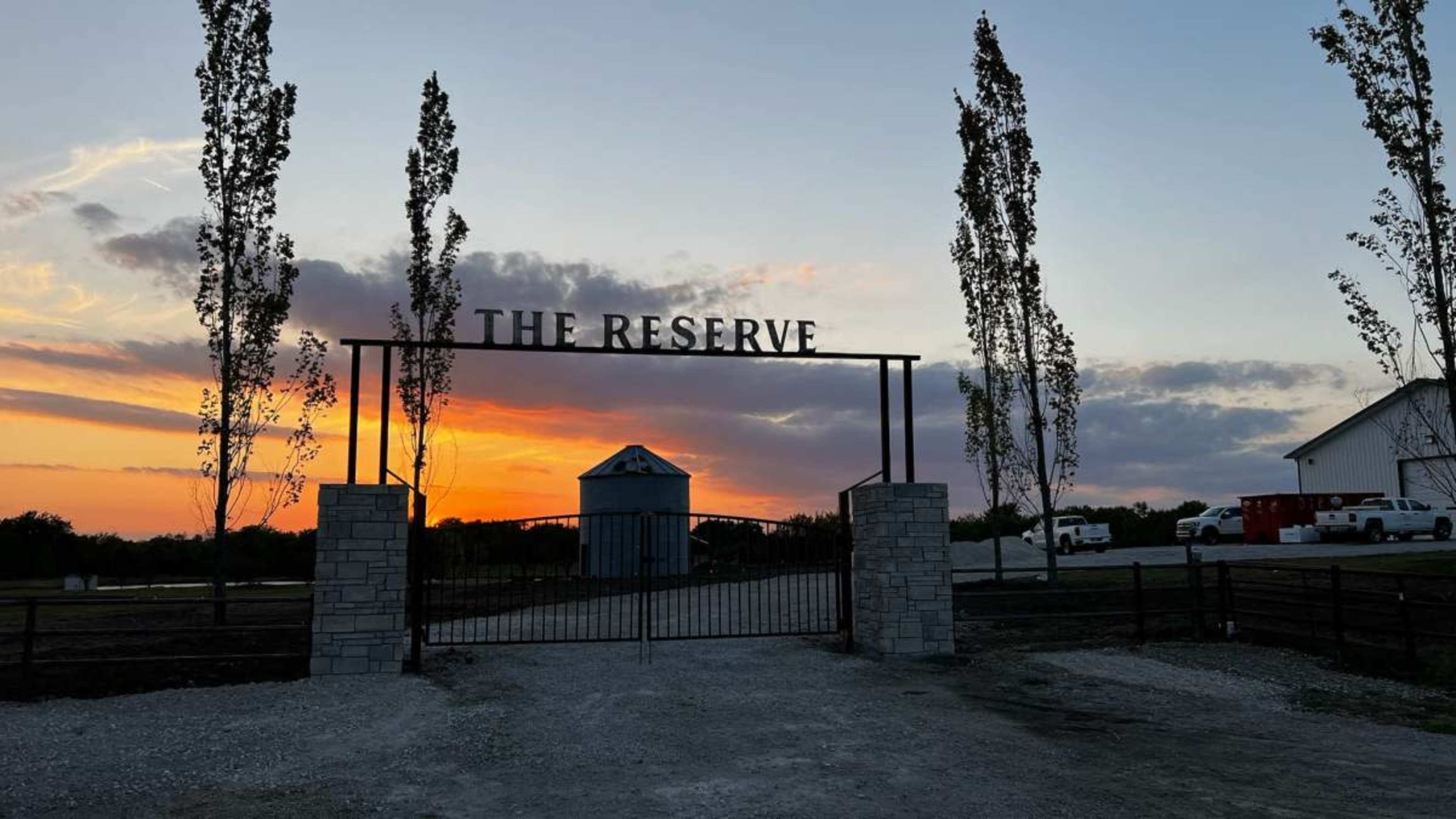 The entrance gate of "The Reserve" is framed by tall trees against a sunset sky, with a gravel driveway leading towards a building and vehicles in the background.