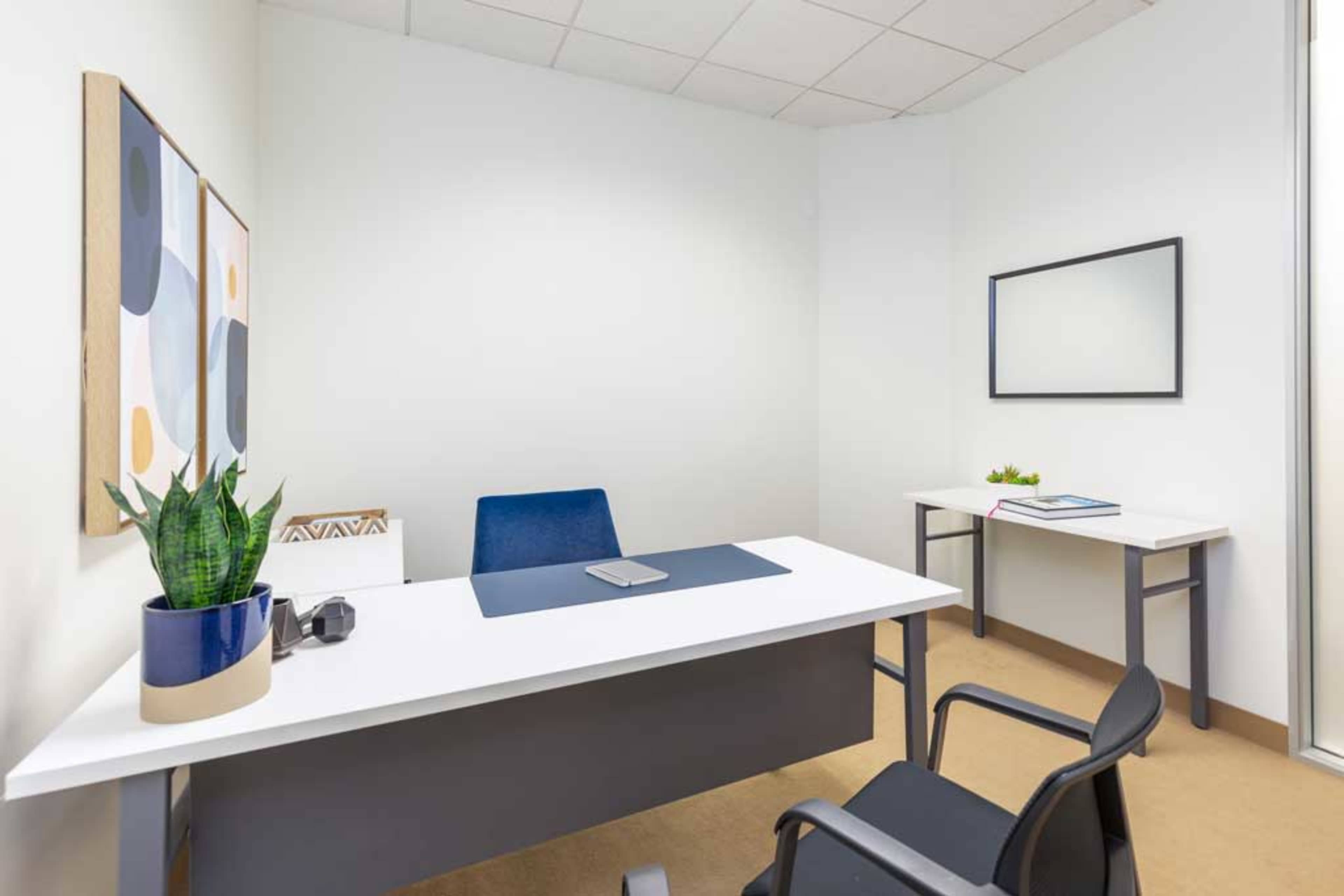 A small office space features a desk with a plant, a chair, and a side table against white walls adorned with abstract art.