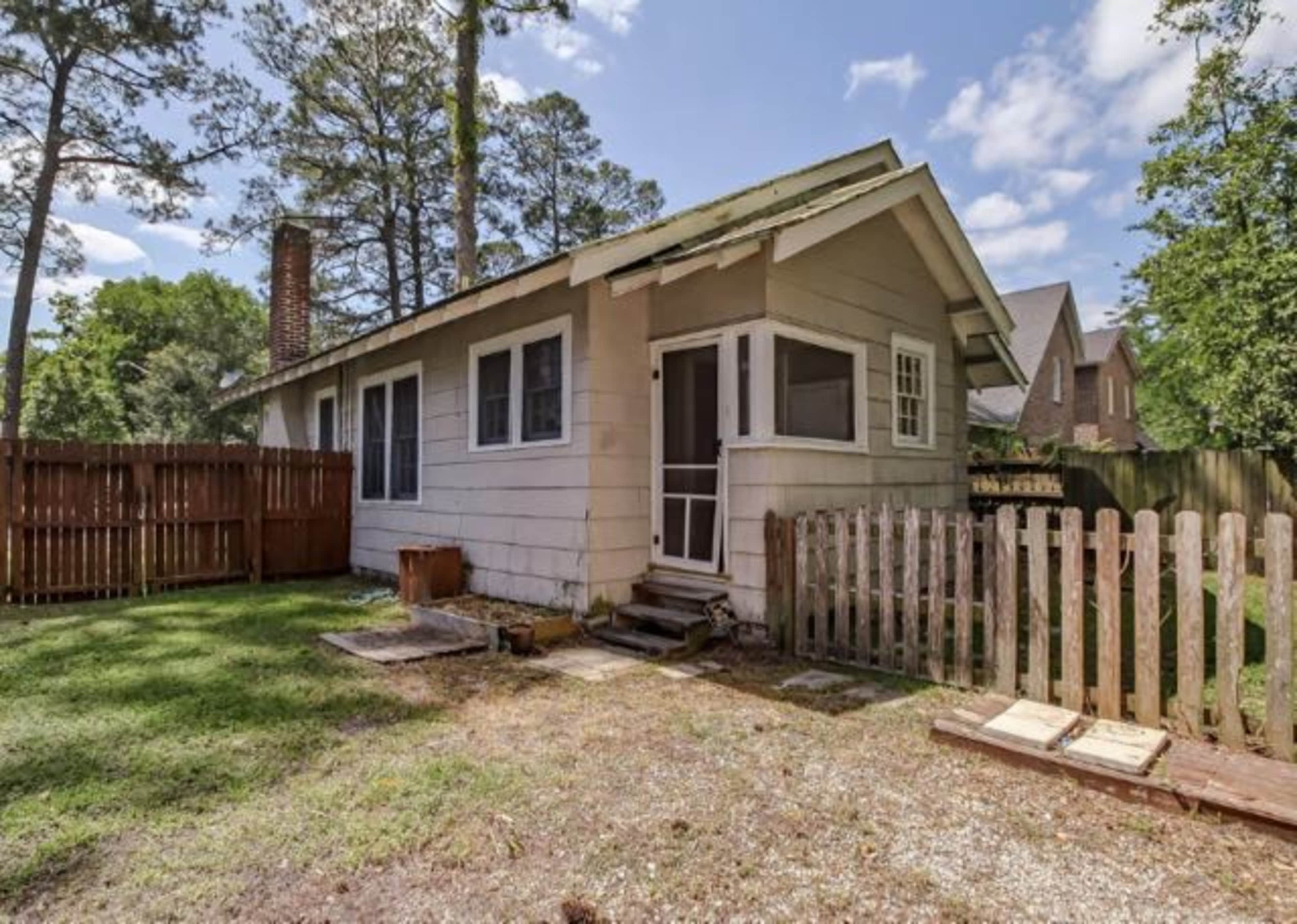 A small, single-story house with a front porch and a fenced yard surrounded by trees.