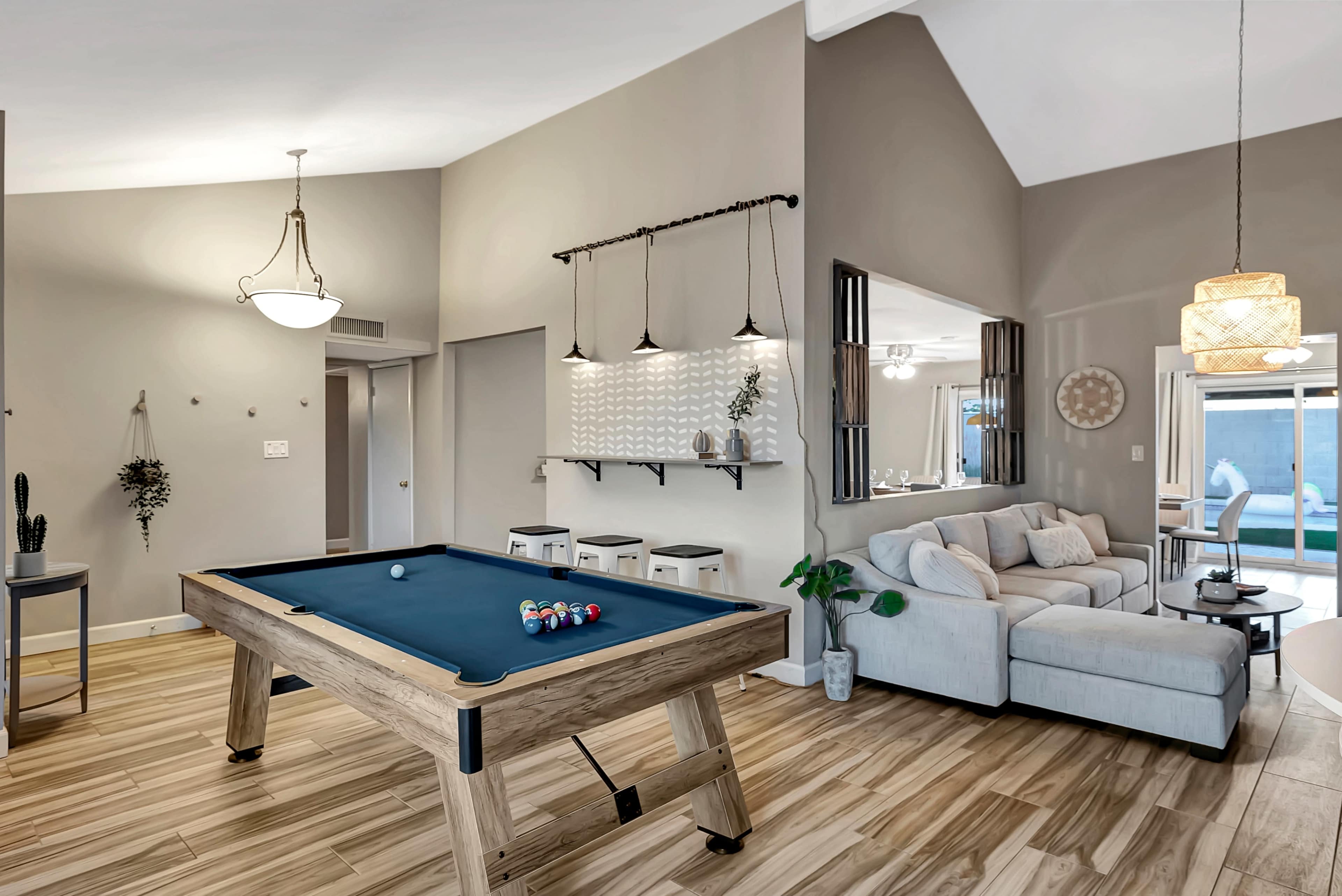 The image shows a modern living space with a pool table, a seating area, and large windows allowing natural light.