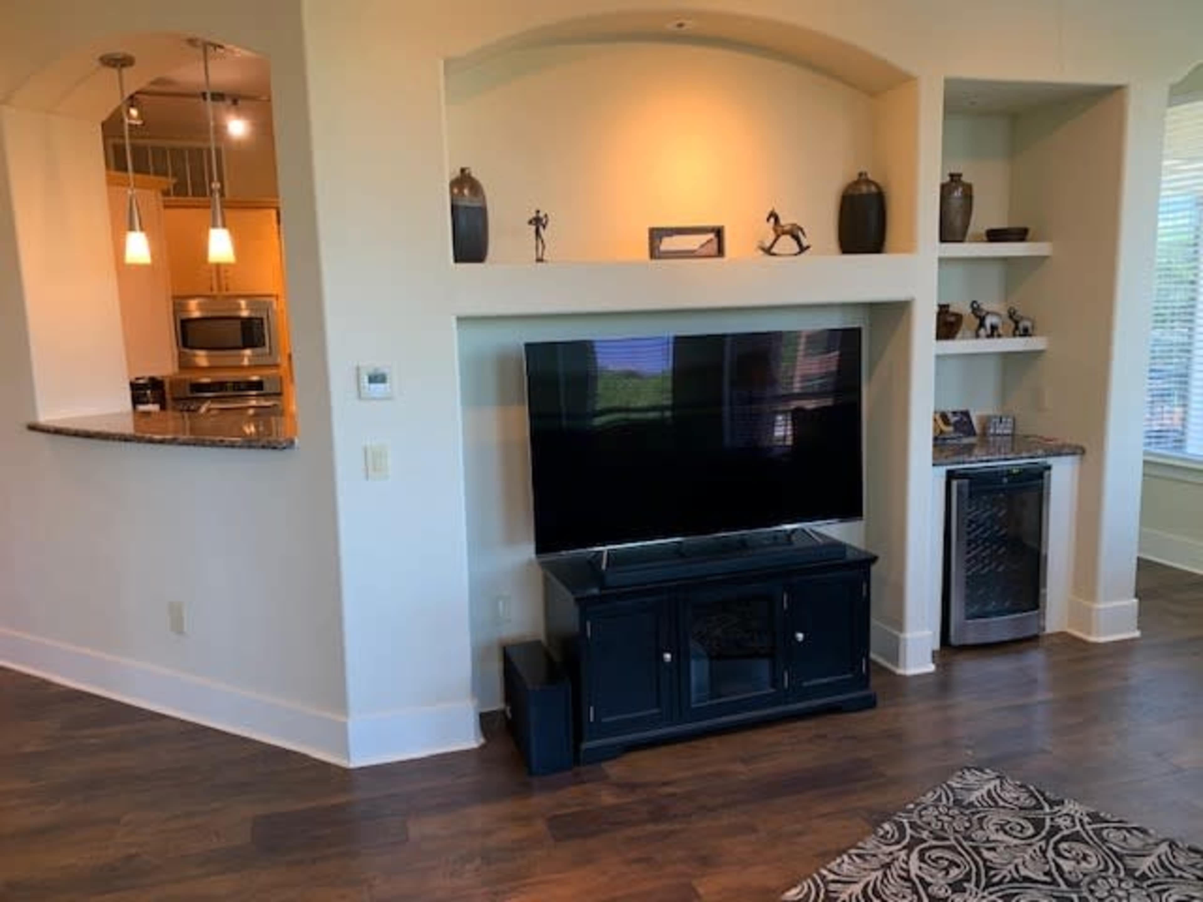 A large television is mounted on a wall unit beside a small refrigerator and decorative items in a well-lit living room.