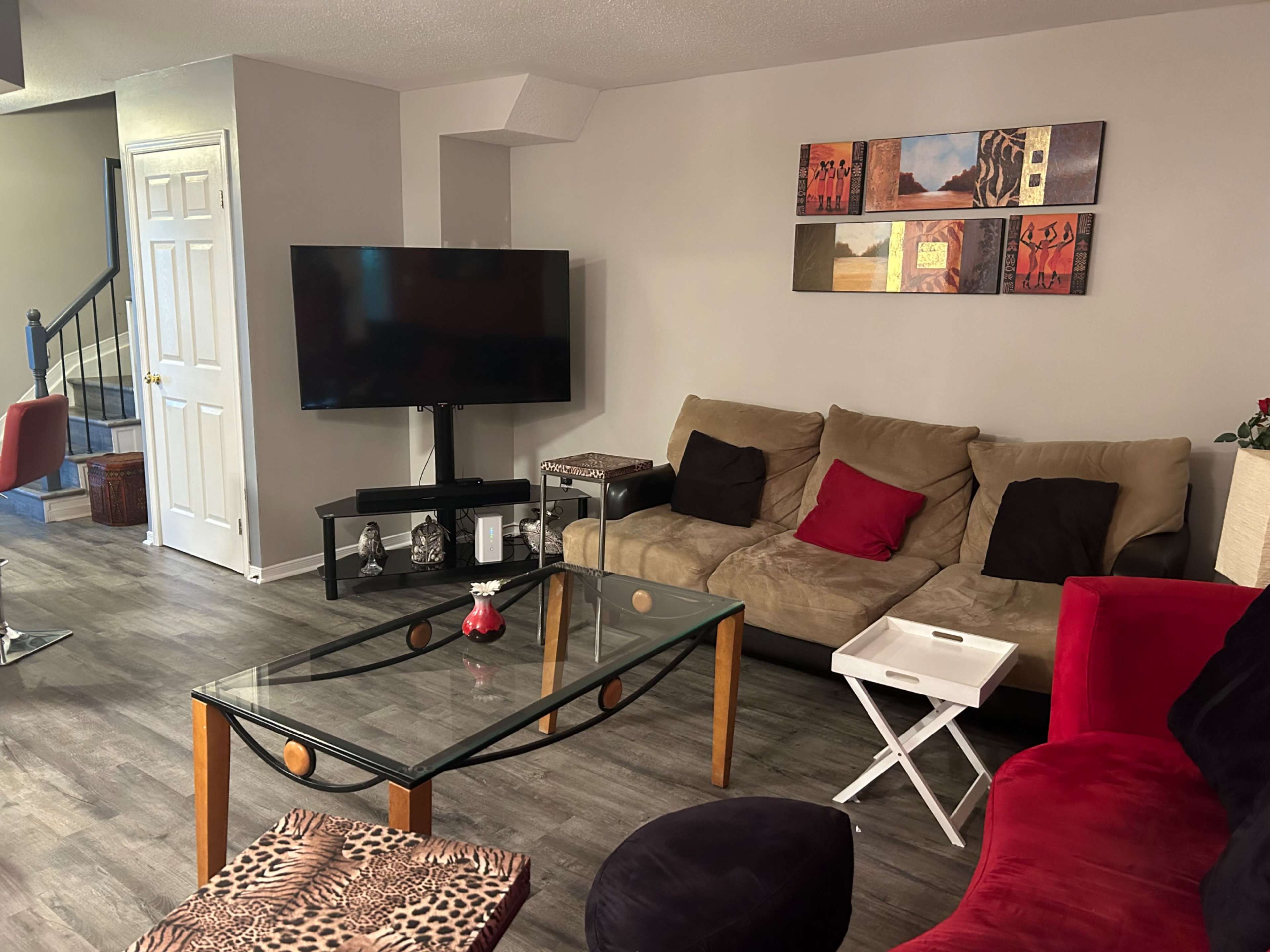 A living room featuring a beige sofa with red and black cushions, a glass coffee table, a television mounted on the wall, and artwork displayed above the couch.