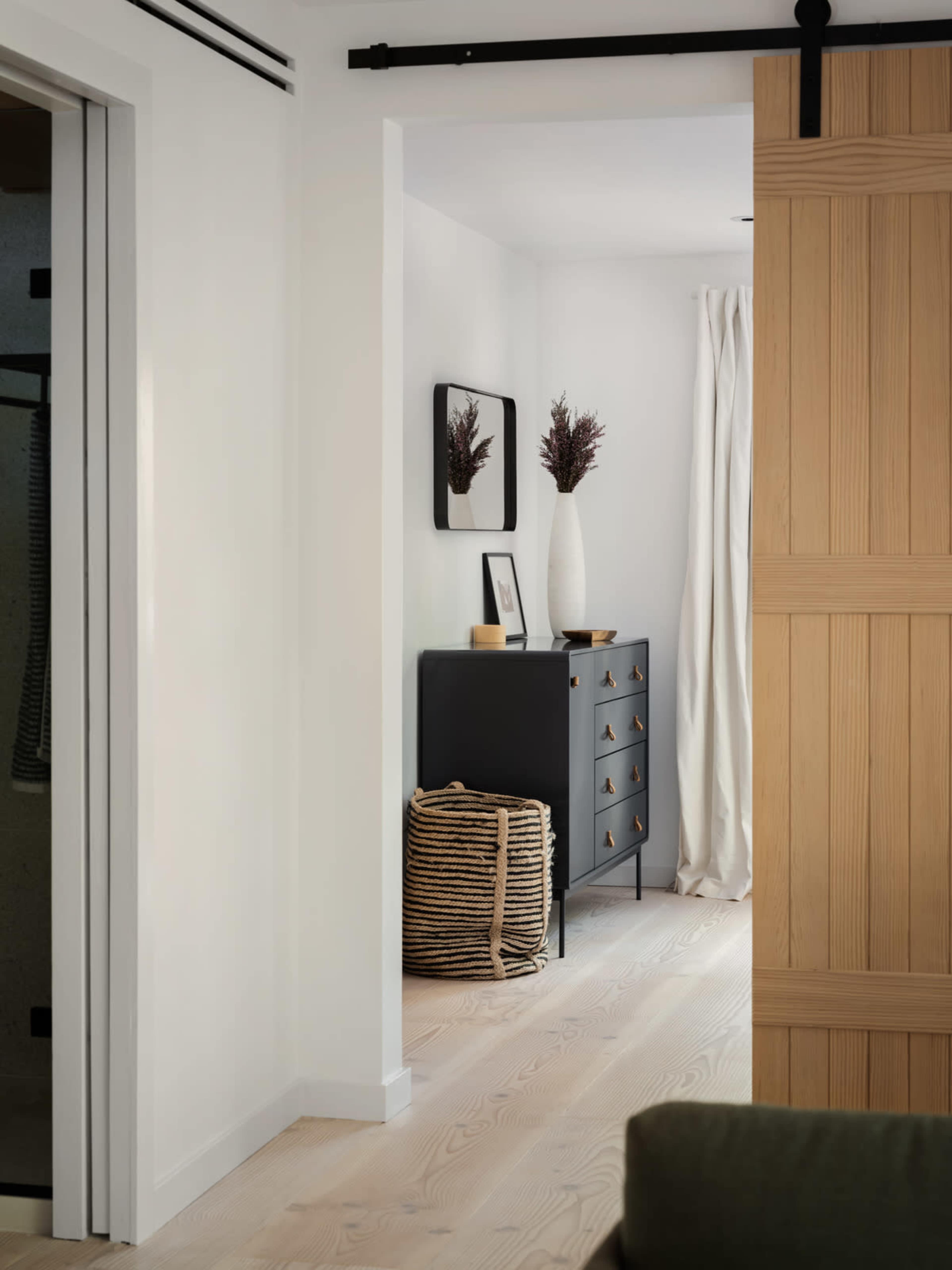 A hallway with a wooden sliding door leads to a room featuring a black dresser, a decorative vase, and a woven basket.