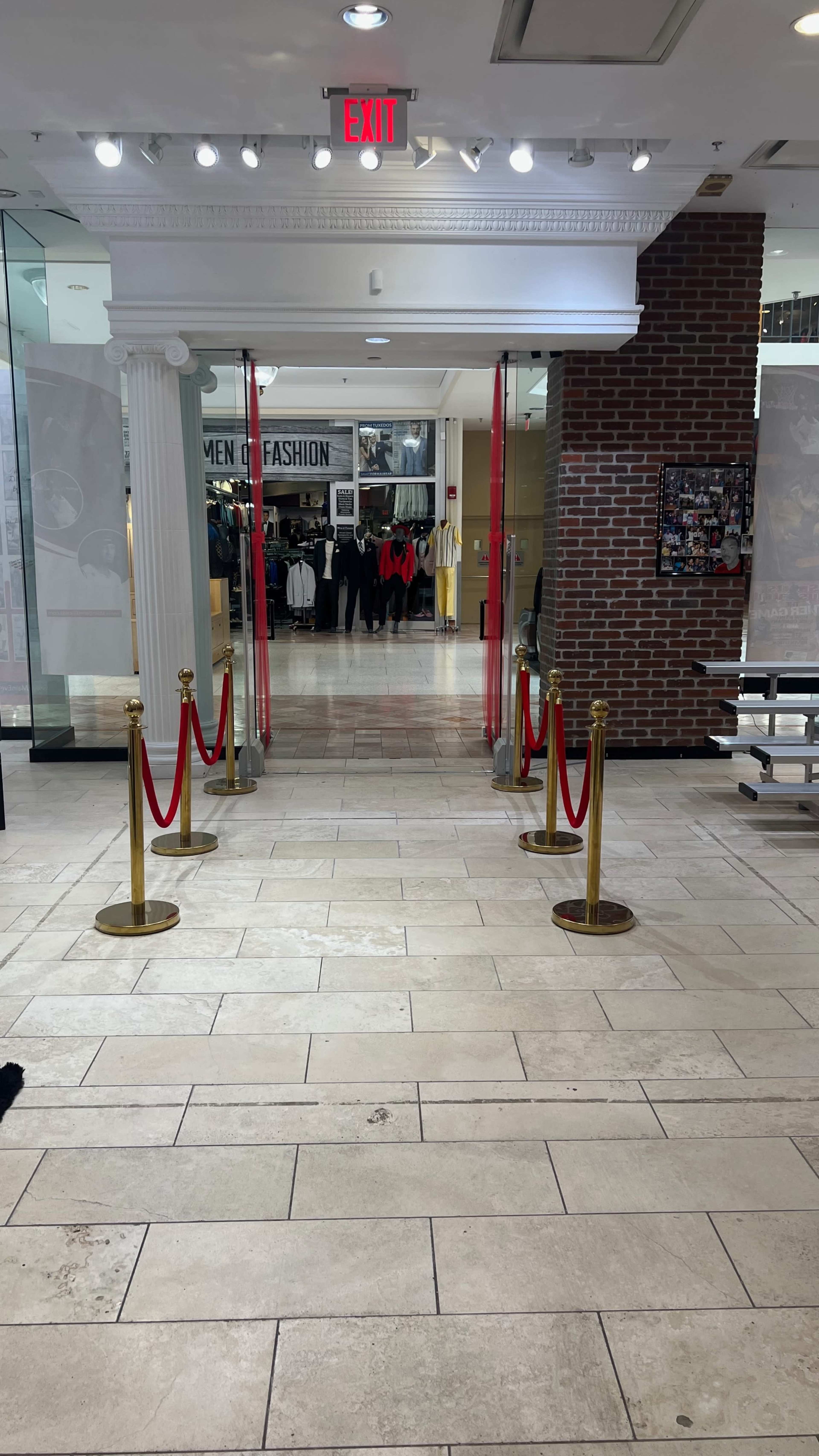 The image shows a mall entrance with red velvet ropes and stanchions leading into a clothing store labeled "MEN FASHION," with shoppers visible in the background.