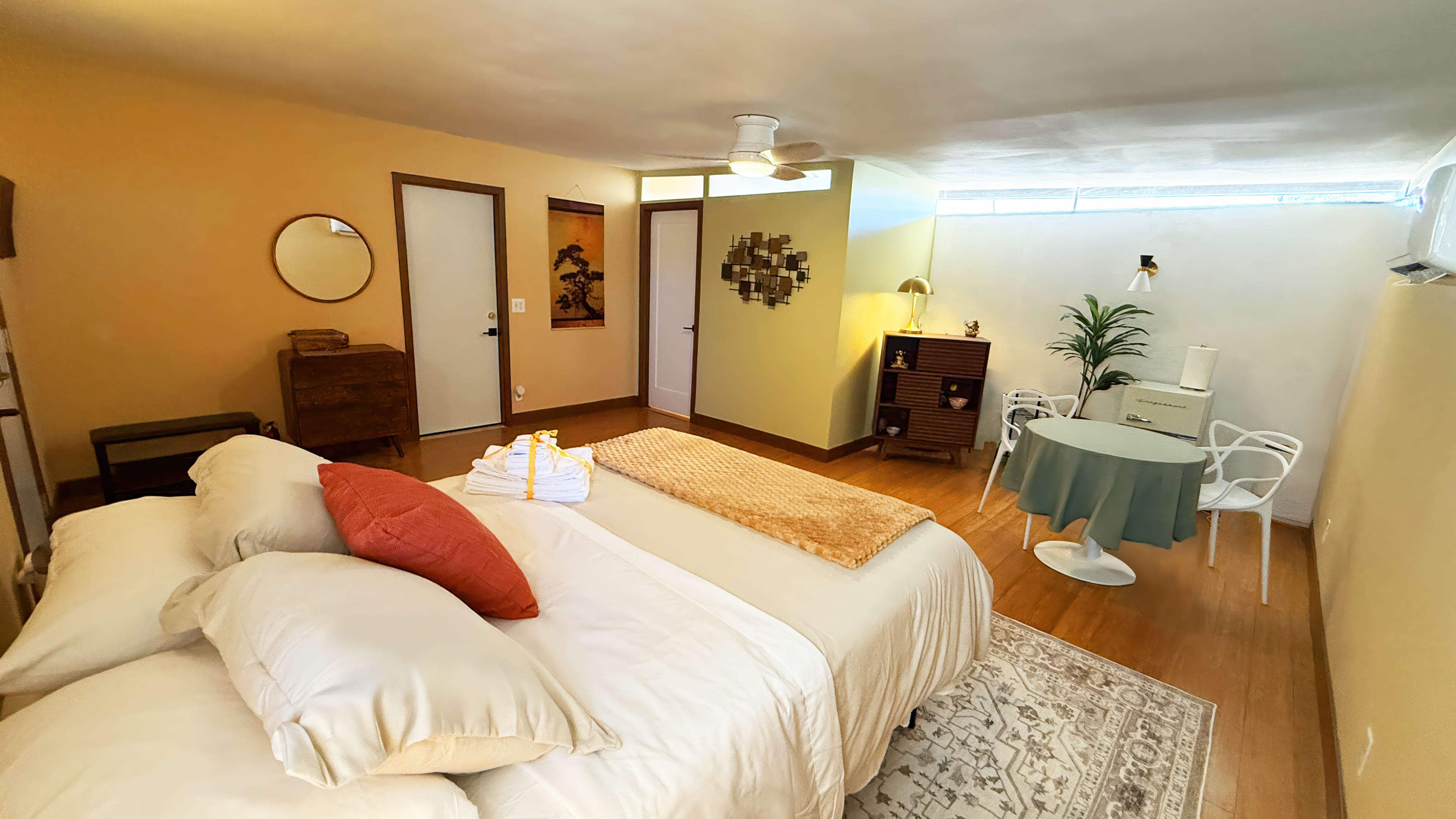 The image shows a bright bedroom with a double bed, a small dining table, and a seating area, featuring wooden furnishings and a fan on the ceiling.