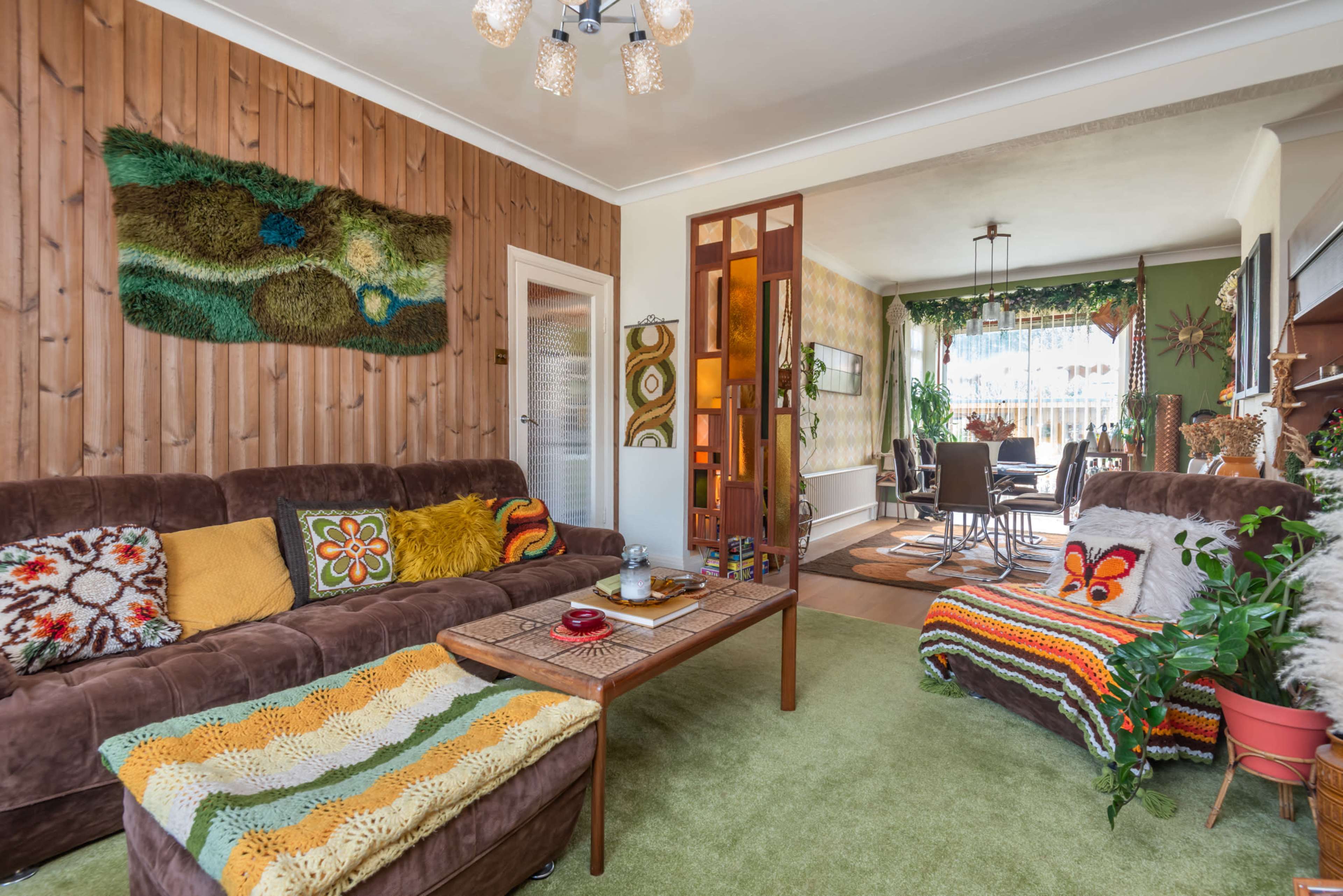 The living room features a brown sofa with colorful crochet blankets, a wooden wall, and a divider leading to a dining area filled with plants.