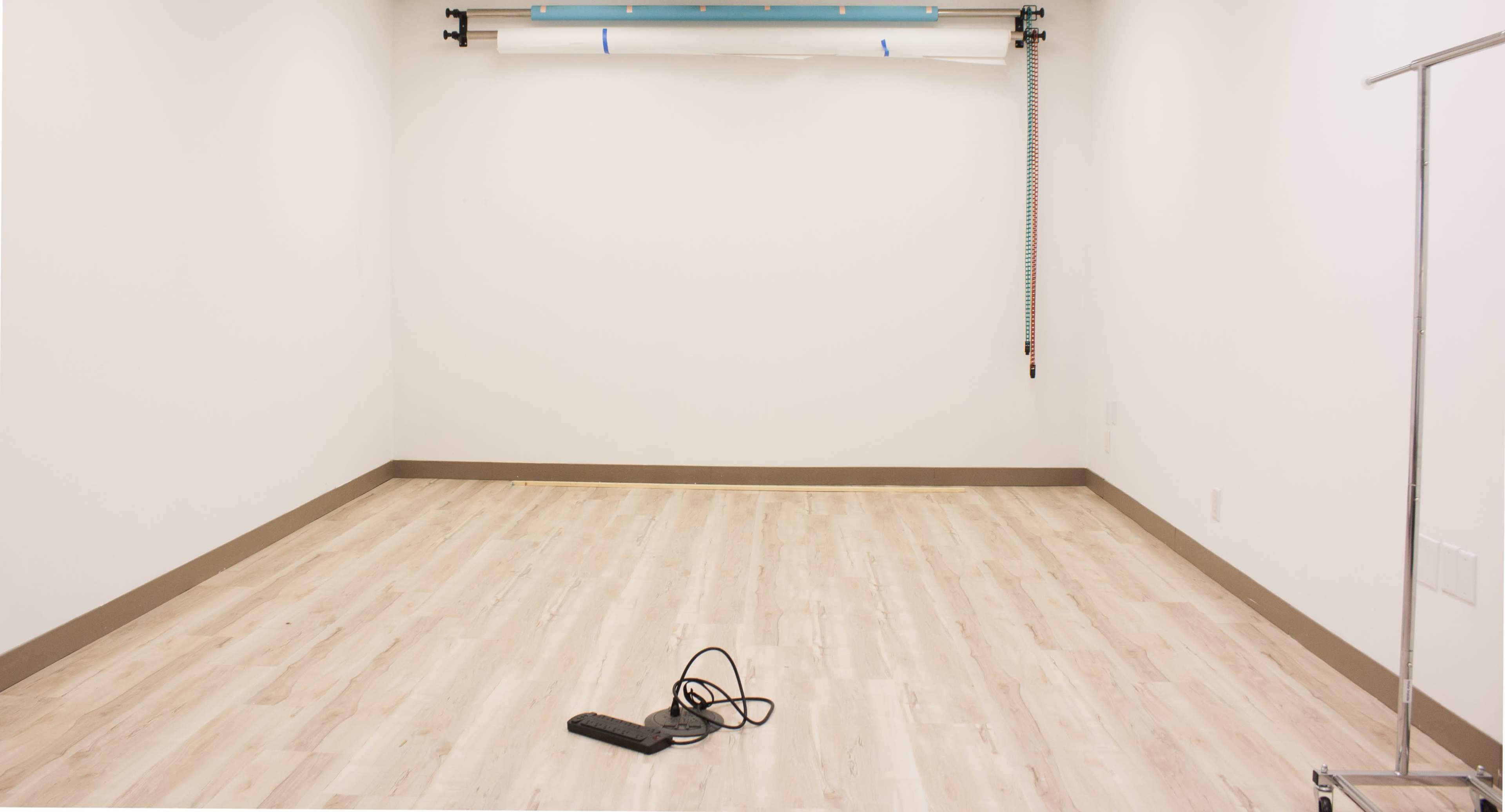 The image shows an empty room with light-colored flooring and a blank white wall, featuring a power strip on the floor.