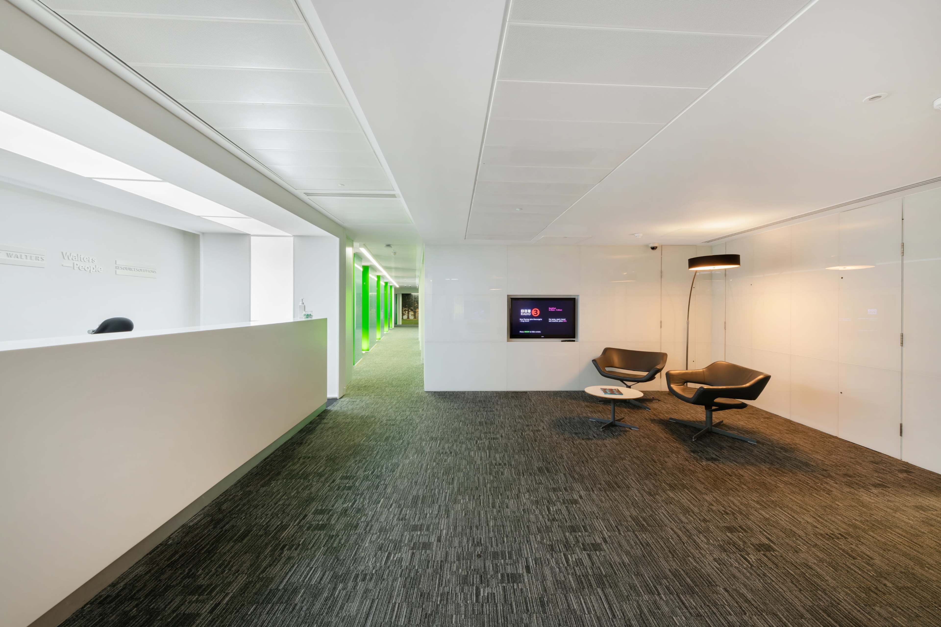 The image shows a modern office reception area with a minimalist design, featuring two chairs and a large digital screen on the wall.