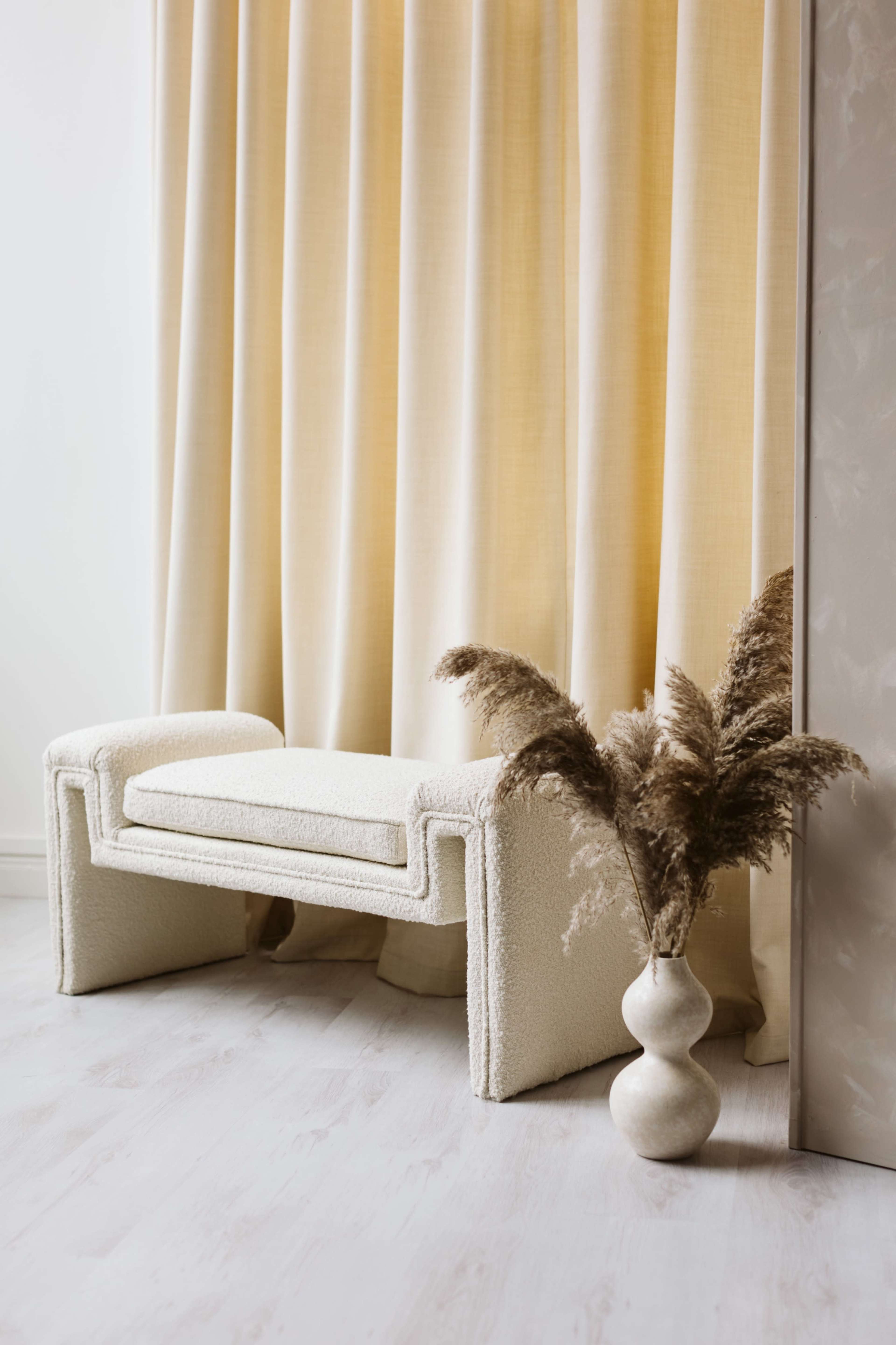 A plush, textured bench sits beside a light yellow curtain, with a vase holding dried pampas grass positioned nearby.