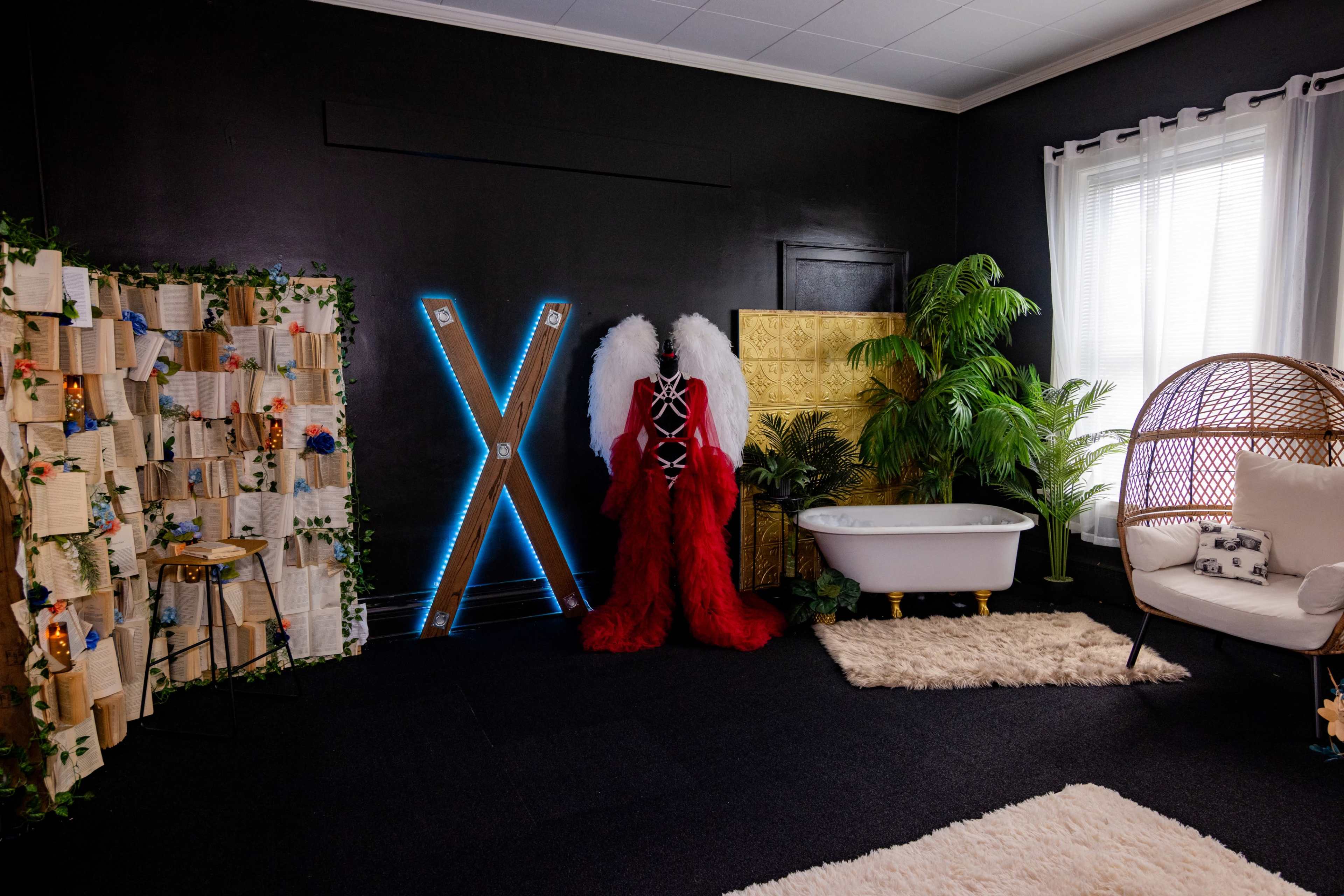 The room features a black wall, a clawfoot bathtub, a large decorative "X" in blue lights, a wall of books, a rattan chair, and various plants.