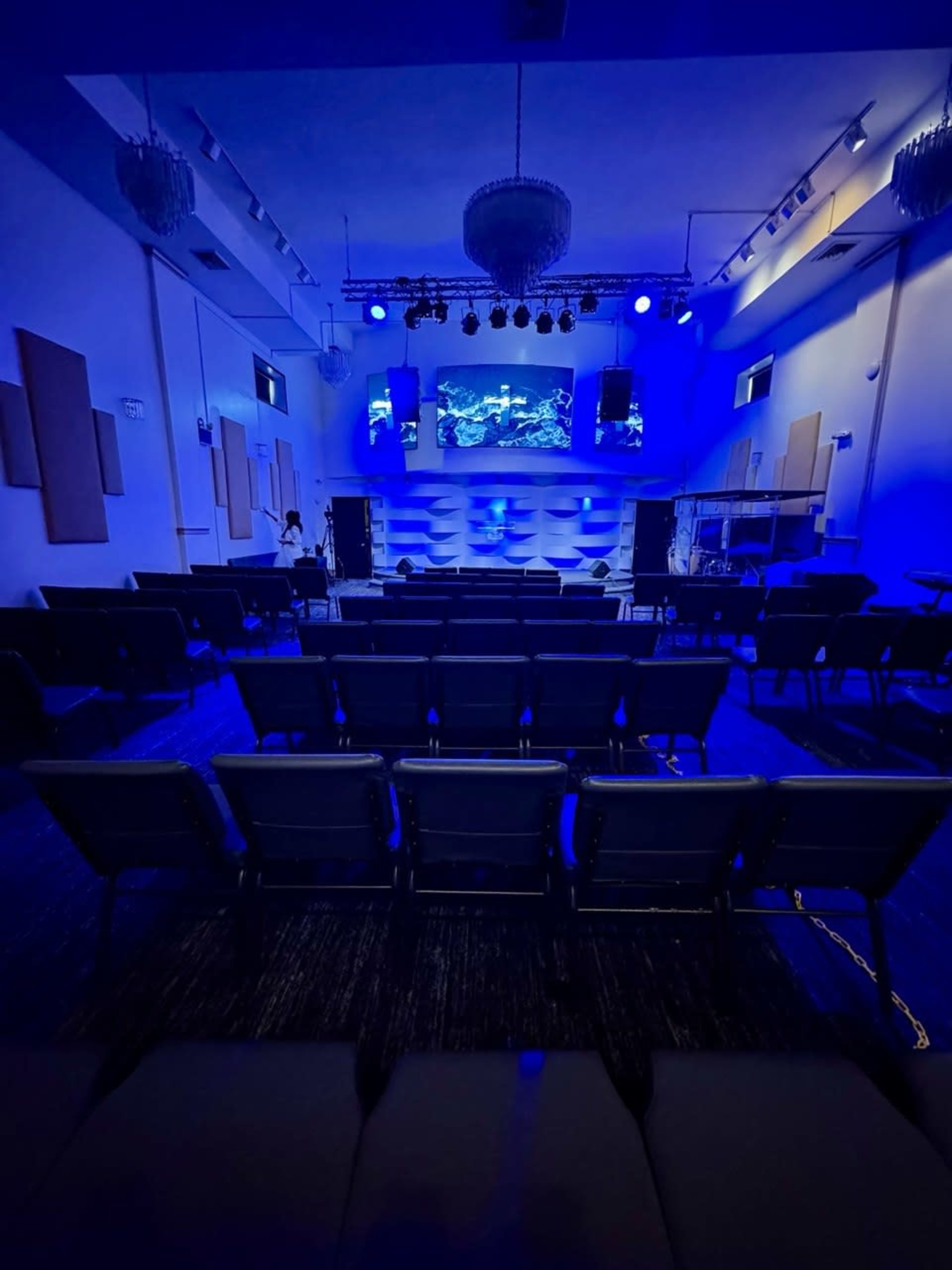 The image shows an empty auditorium with rows of black chairs, blue lighting, and a large screen displaying visuals at the front.