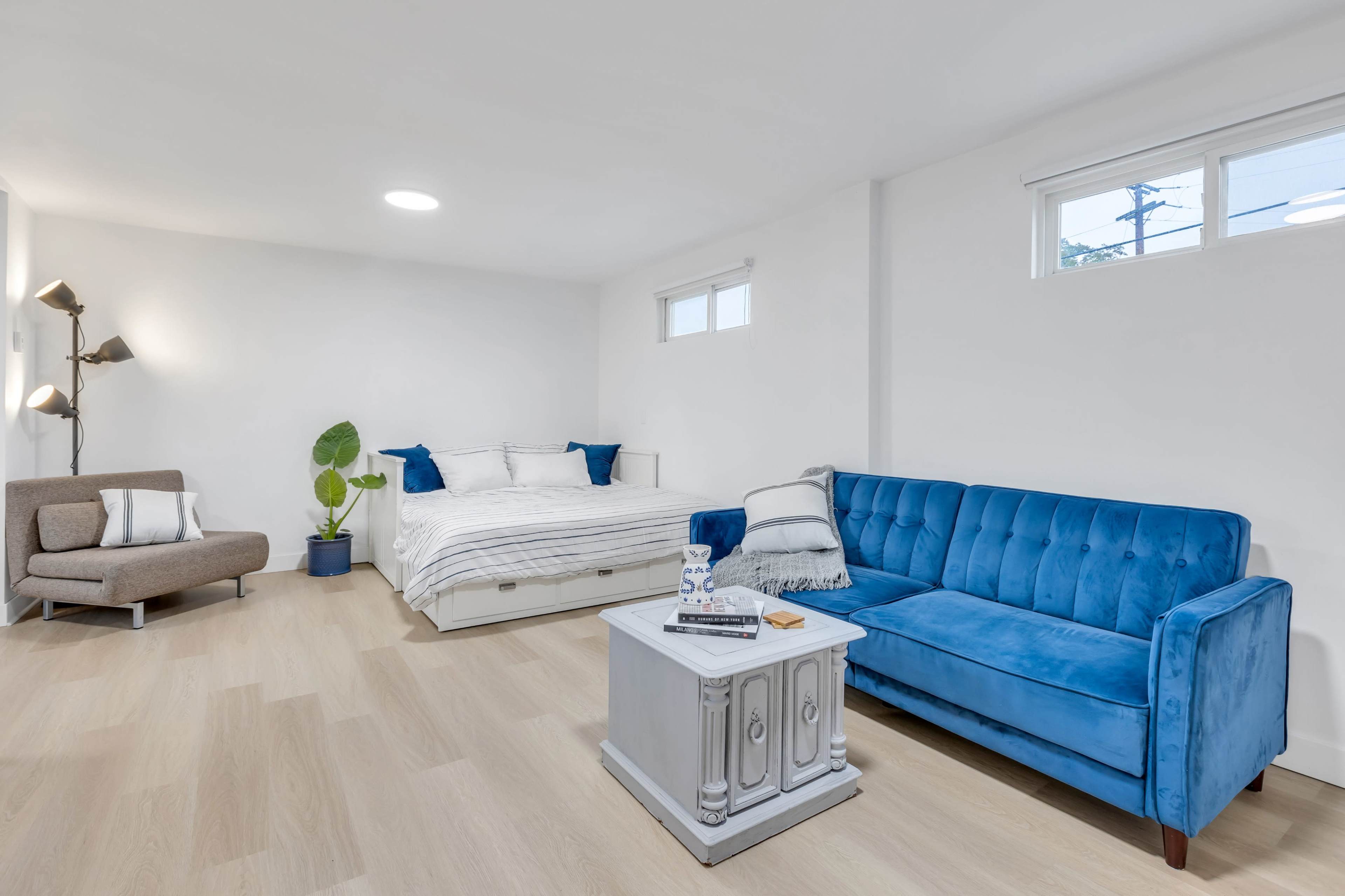 A modern, minimalistic living space featuring a blue couch, a light-colored bed, a small side table, and a potted plant.