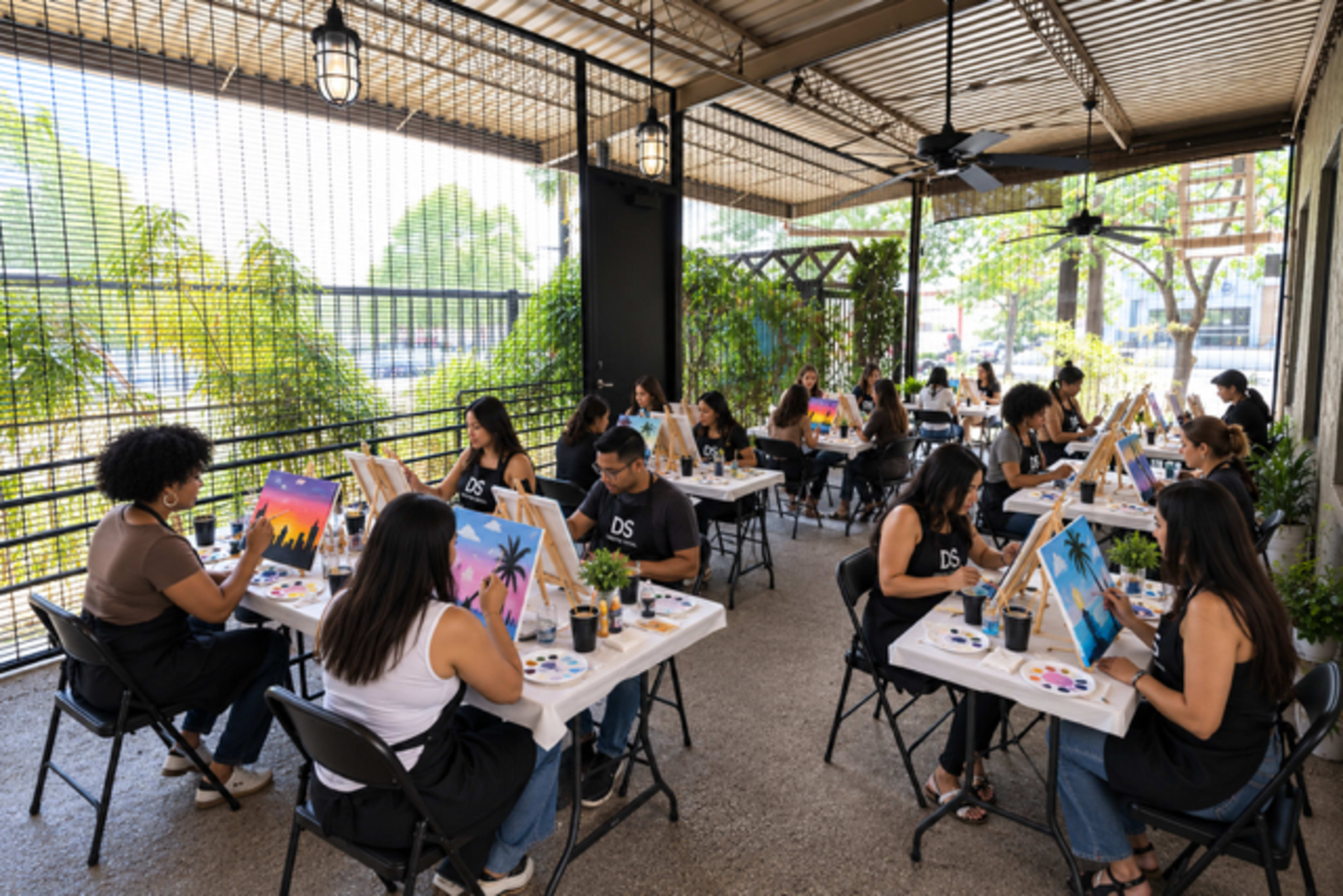 A group of people paint on canvases at tables in an outdoor space surrounded by greenery.