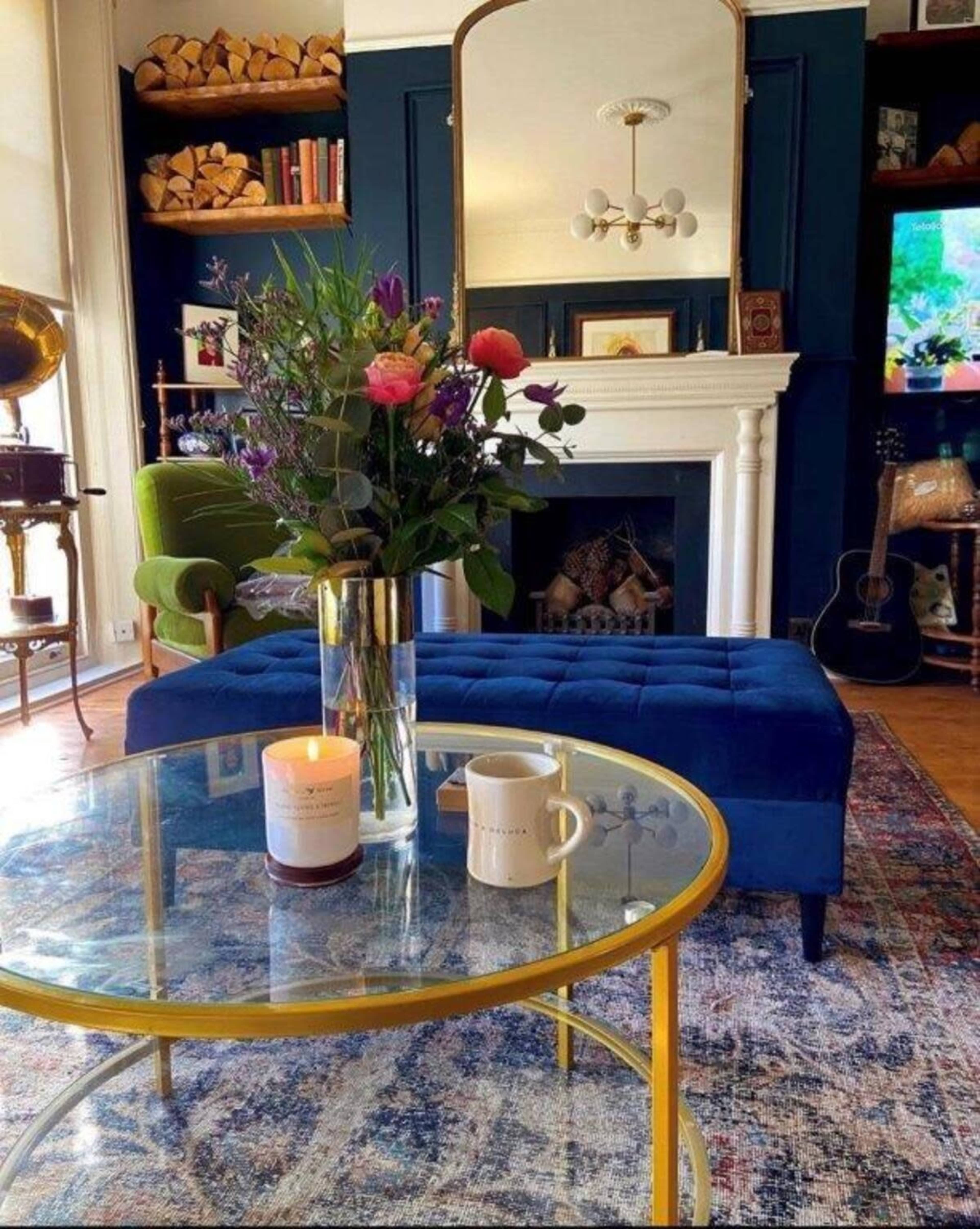 A cozy living room with a colorful vase of flowers on a glass coffee table, a blue tufted ottoman, and a fireplace surrounded by decorative items.