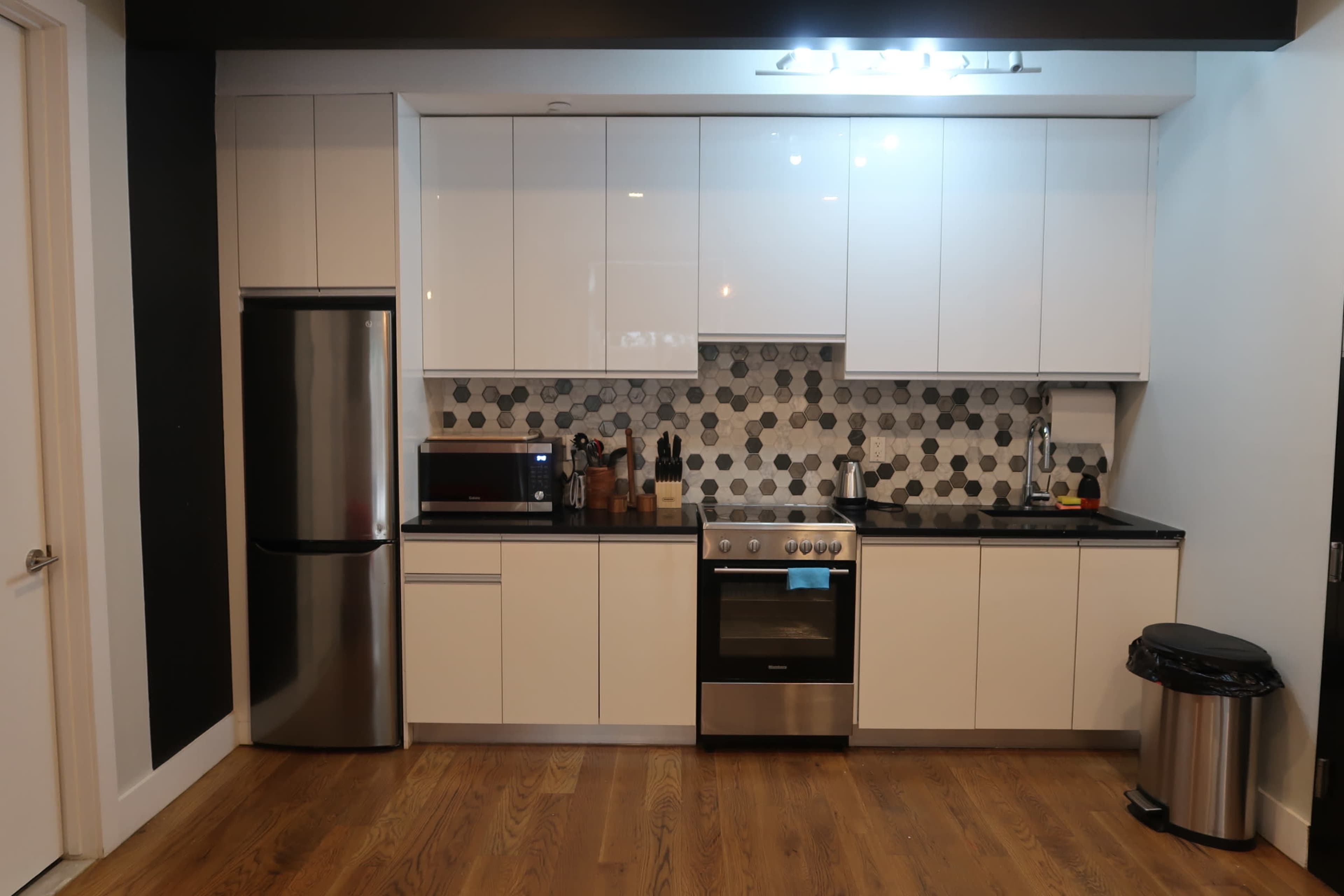 A modern kitchen featuring a stainless steel refrigerator, a black countertop with a stove, an oven, a microwave, and a trash bin, all arranged against a wall with hexagonal tiles.