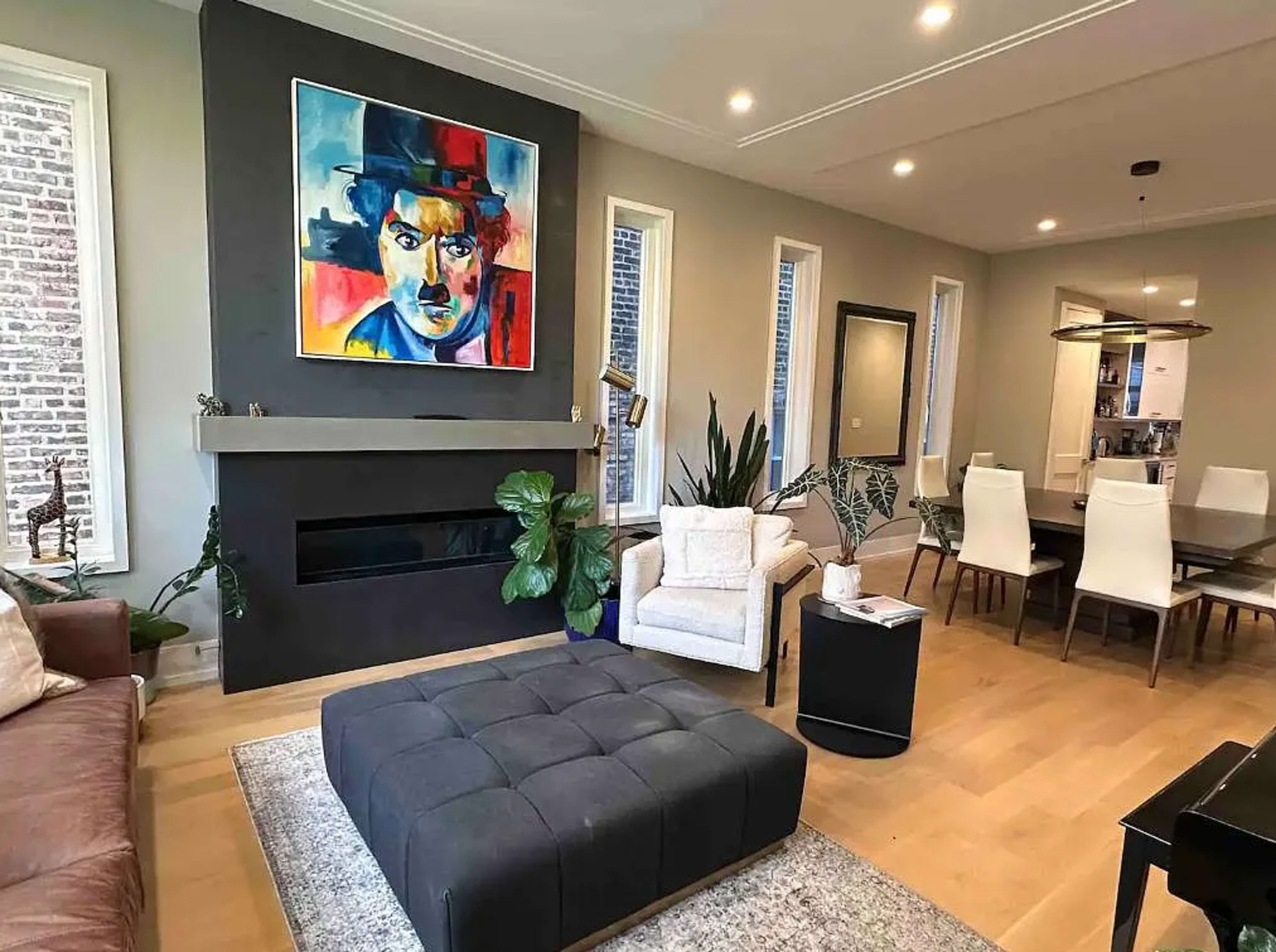 The image shows a modern living room with a large painting on the wall, a sectional sofa, an ottoman, indoor plants, and a dining area visible in the background.