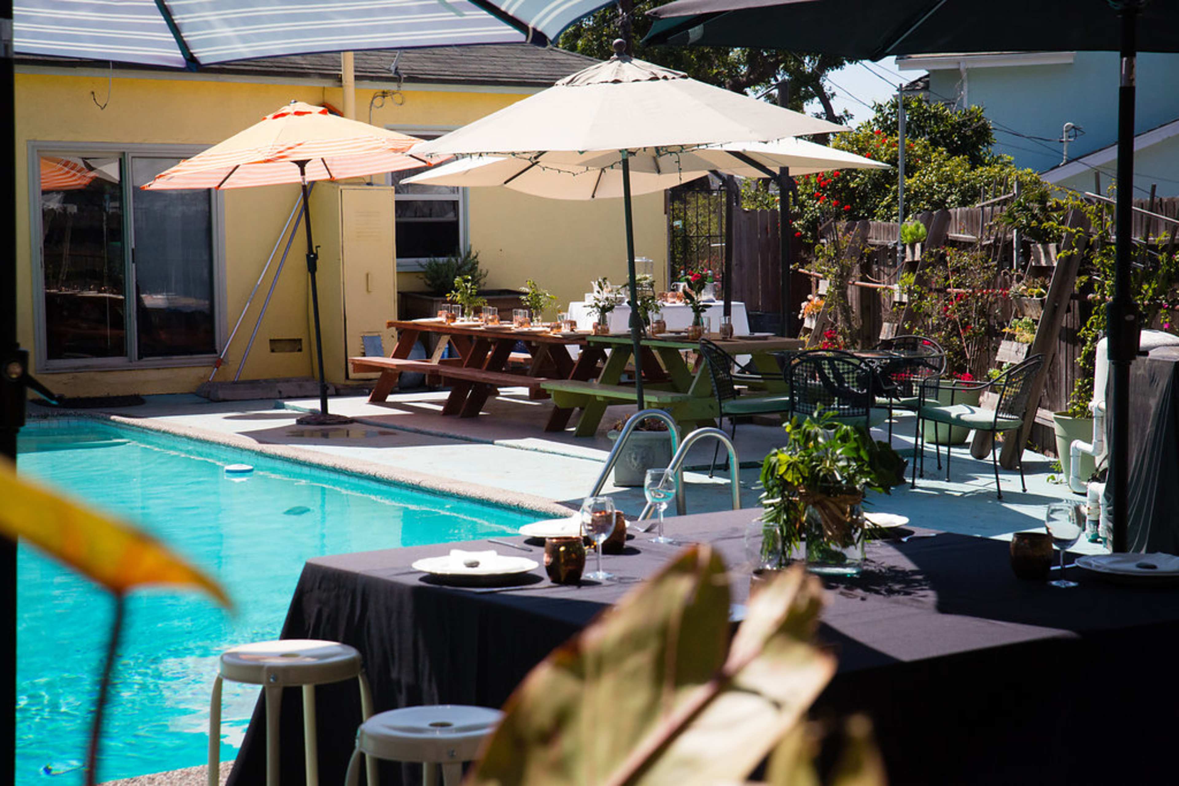 The image shows a backyard scene with a swimming pool, dining tables set for a meal, and various umbrellas providing shade.