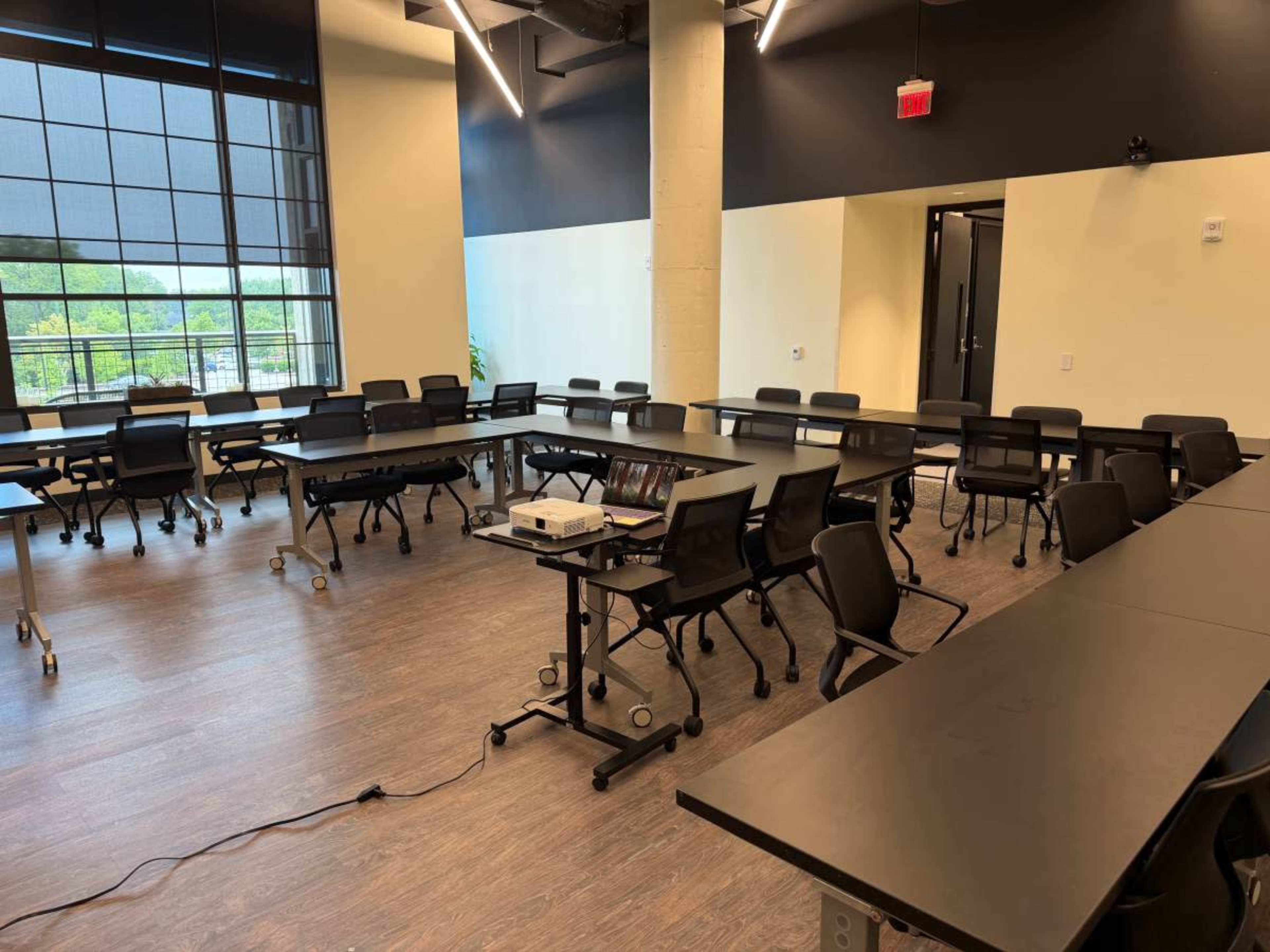 The image shows a conference room with several rows of tables and chairs arranged for a meeting, along with a projector on a stand.