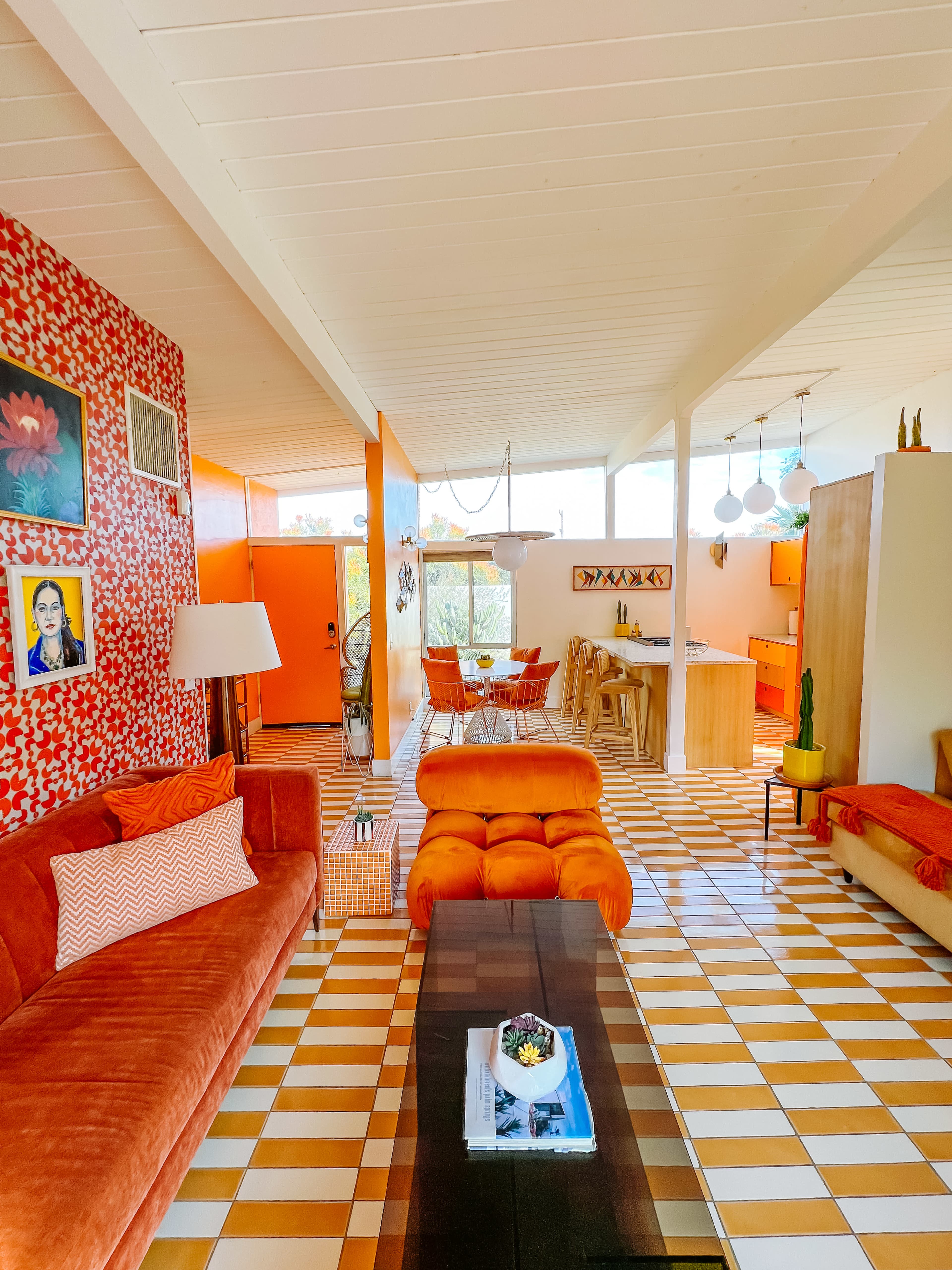 The image depicts a brightly colored mid-century modern living space featuring orange sofas, patterned walls, and a tiled floor, with a dining area and kitchen in the background.