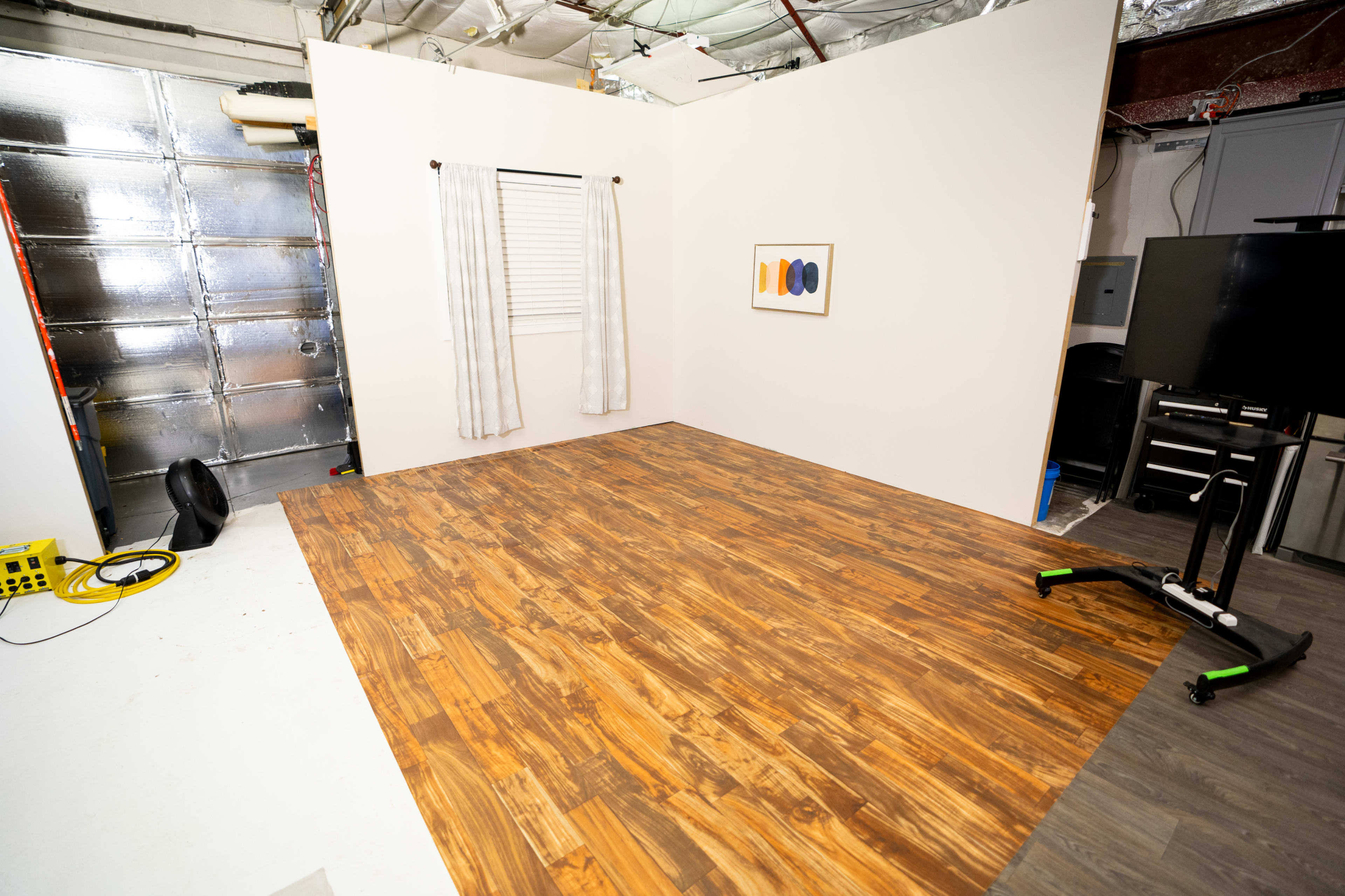 The image depicts a sparsely furnished room with a wood-patterned floor, a window with curtains, and a wall-mounted artwork.