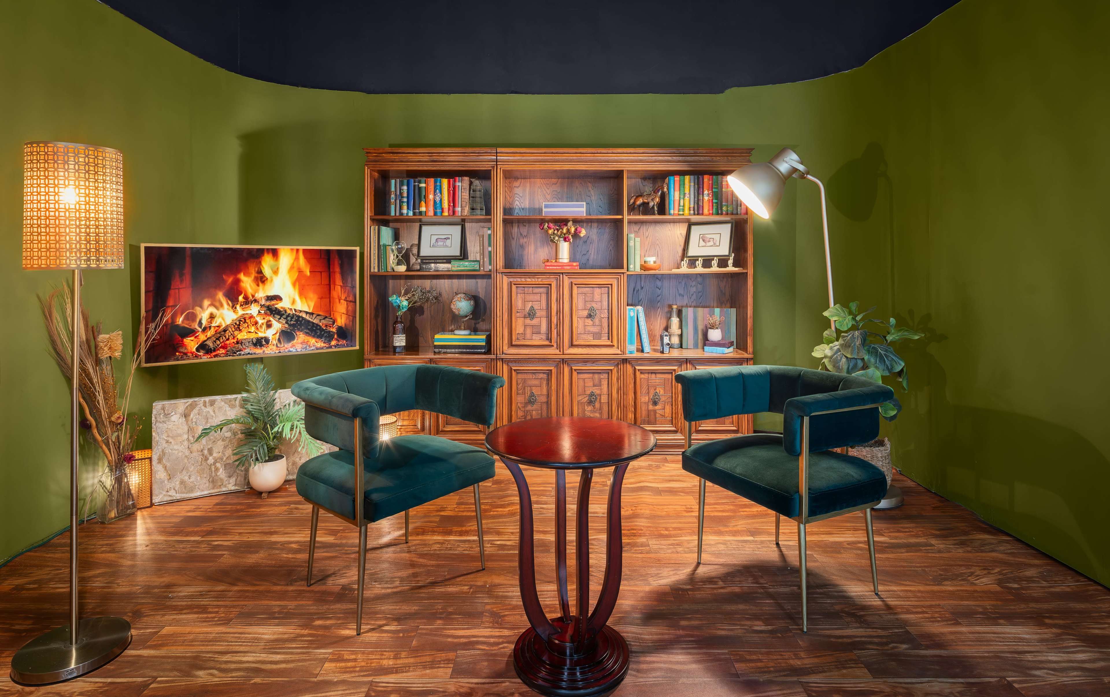 The image features a cozy interior scene with two velvet chairs, a small round table, a bookshelf filled with books, a wall-mounted TV displaying a fireplace, and a standing lamp, all set against a green backdrop.