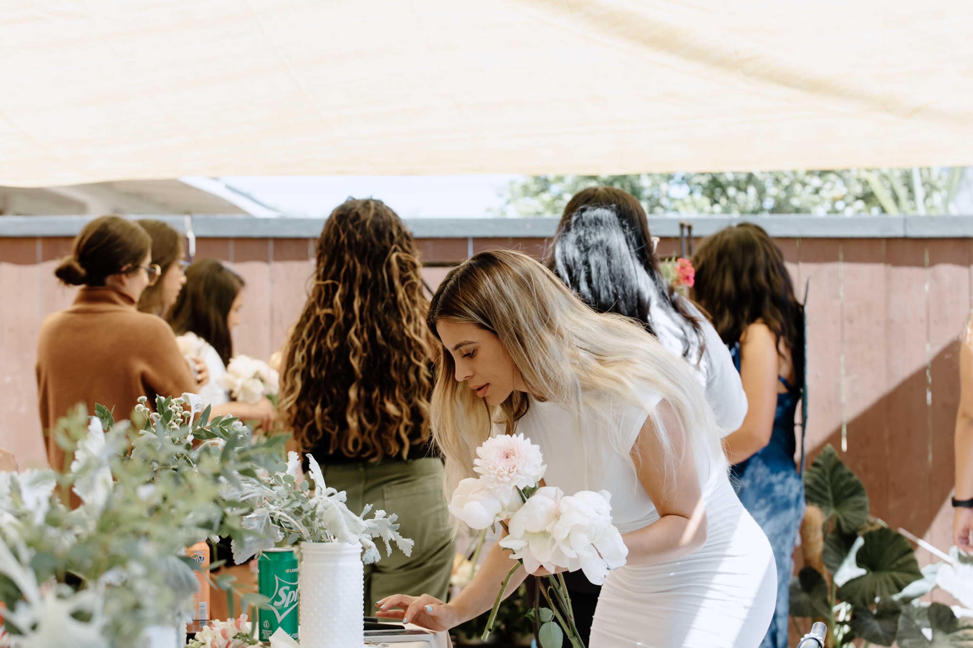 A woman in a white dress examines flowers at a table while a group of people gathers in the background under a canopy.