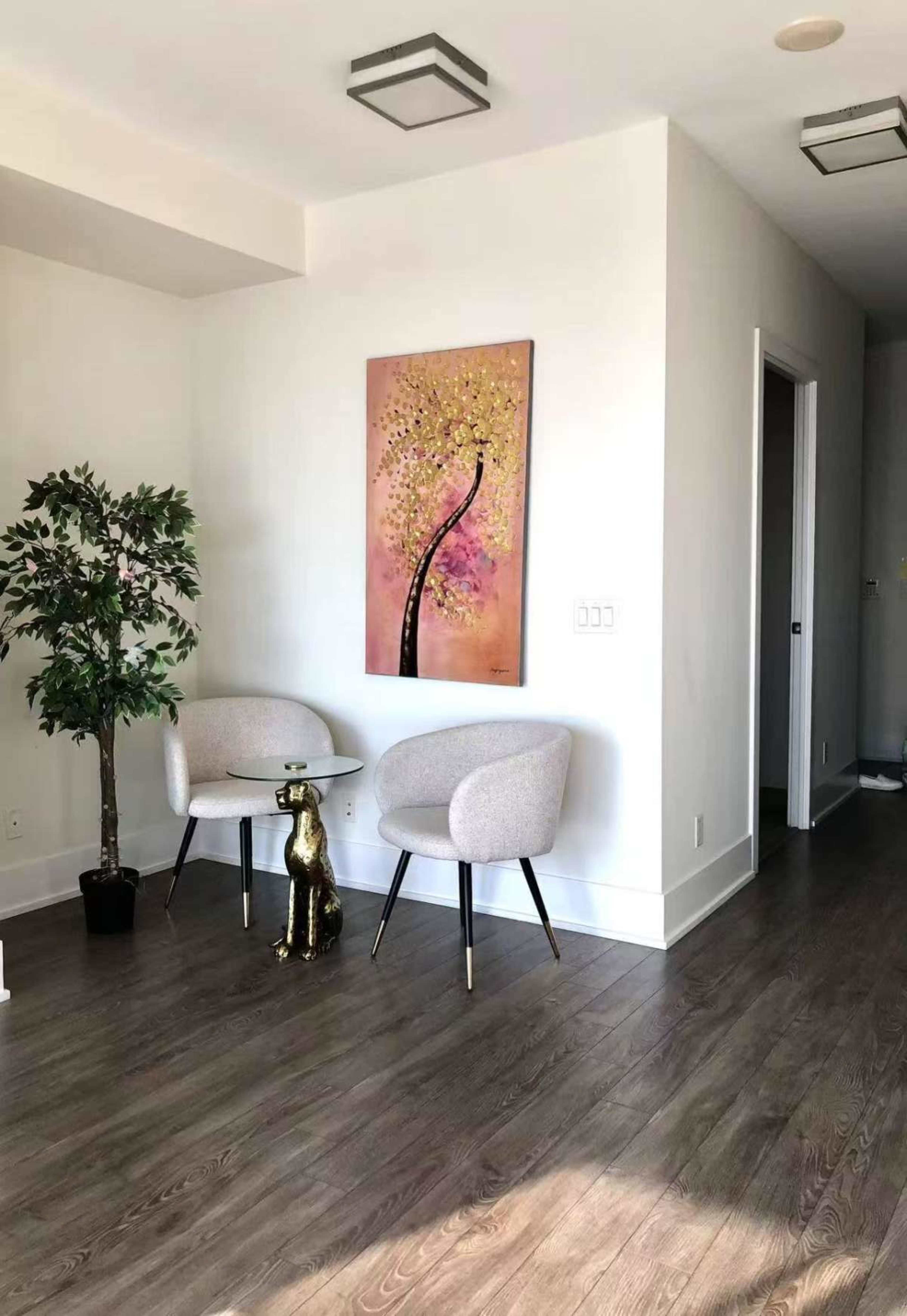 The image shows a small, elegantly furnished corner of a room featuring two light gray chairs, a glass table, a decorative tree, and a painting of a pink tree on the wall.
