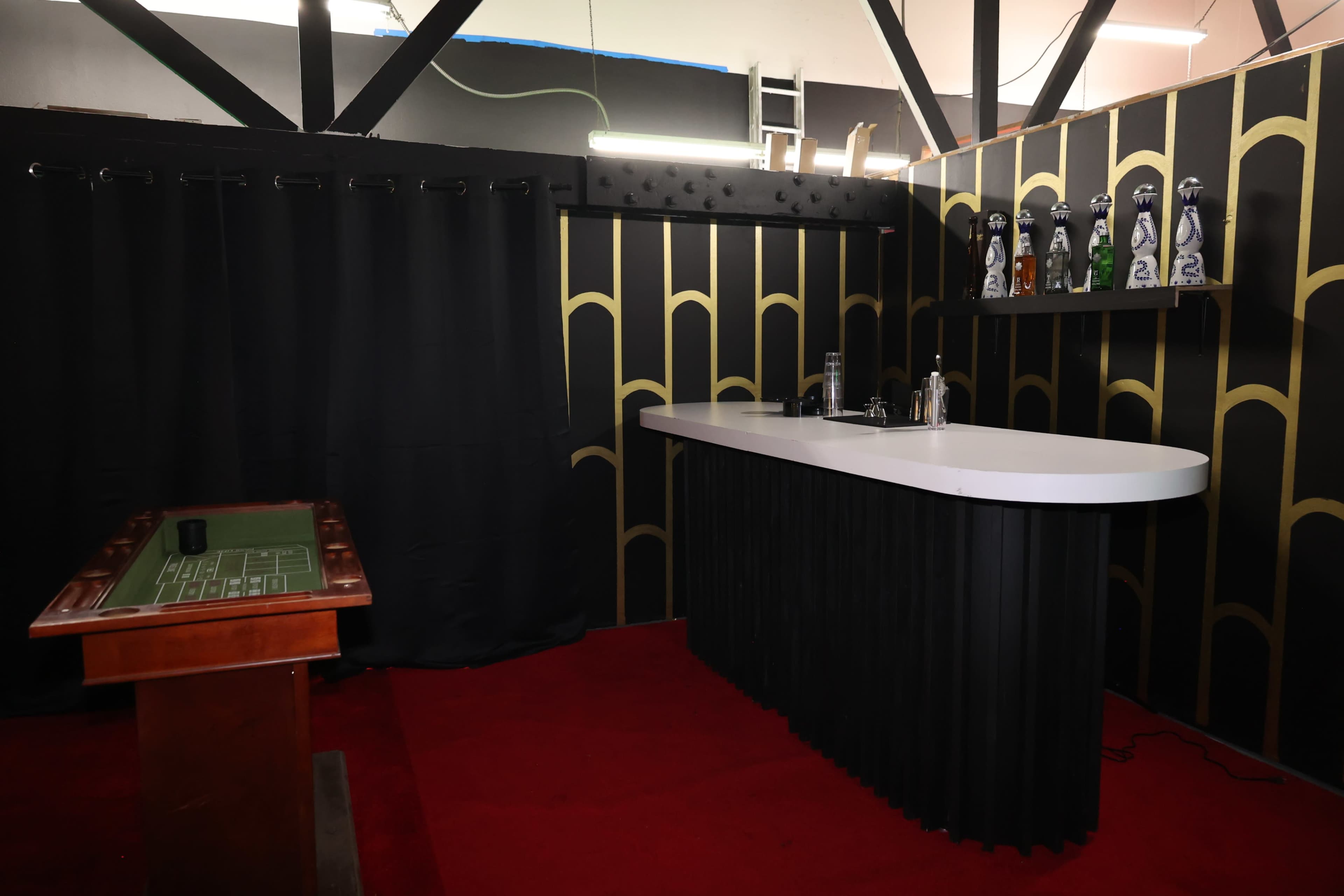 A dimly lit room with a black bar featuring a white countertop, surrounded by dark curtains and patterned walls, alongside a gaming table.