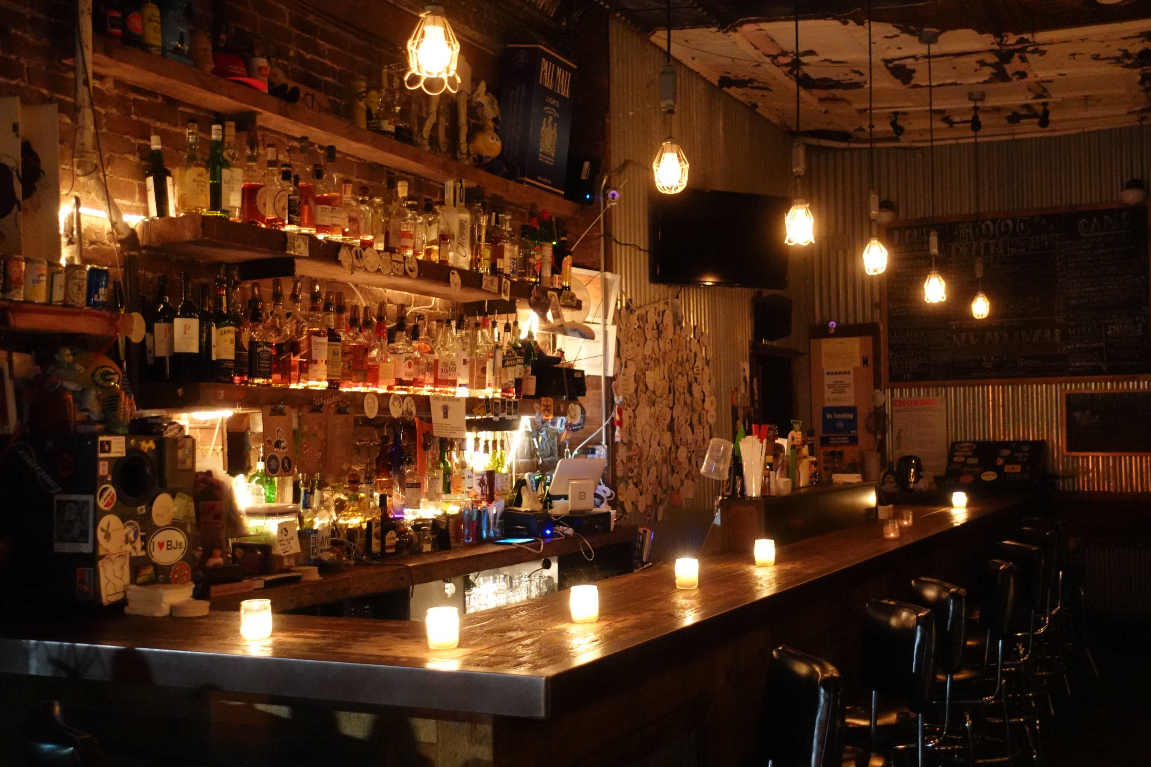 A dimly lit bar with a wooden counter, shelves lined with various bottles, and candlelight illuminating the space.
