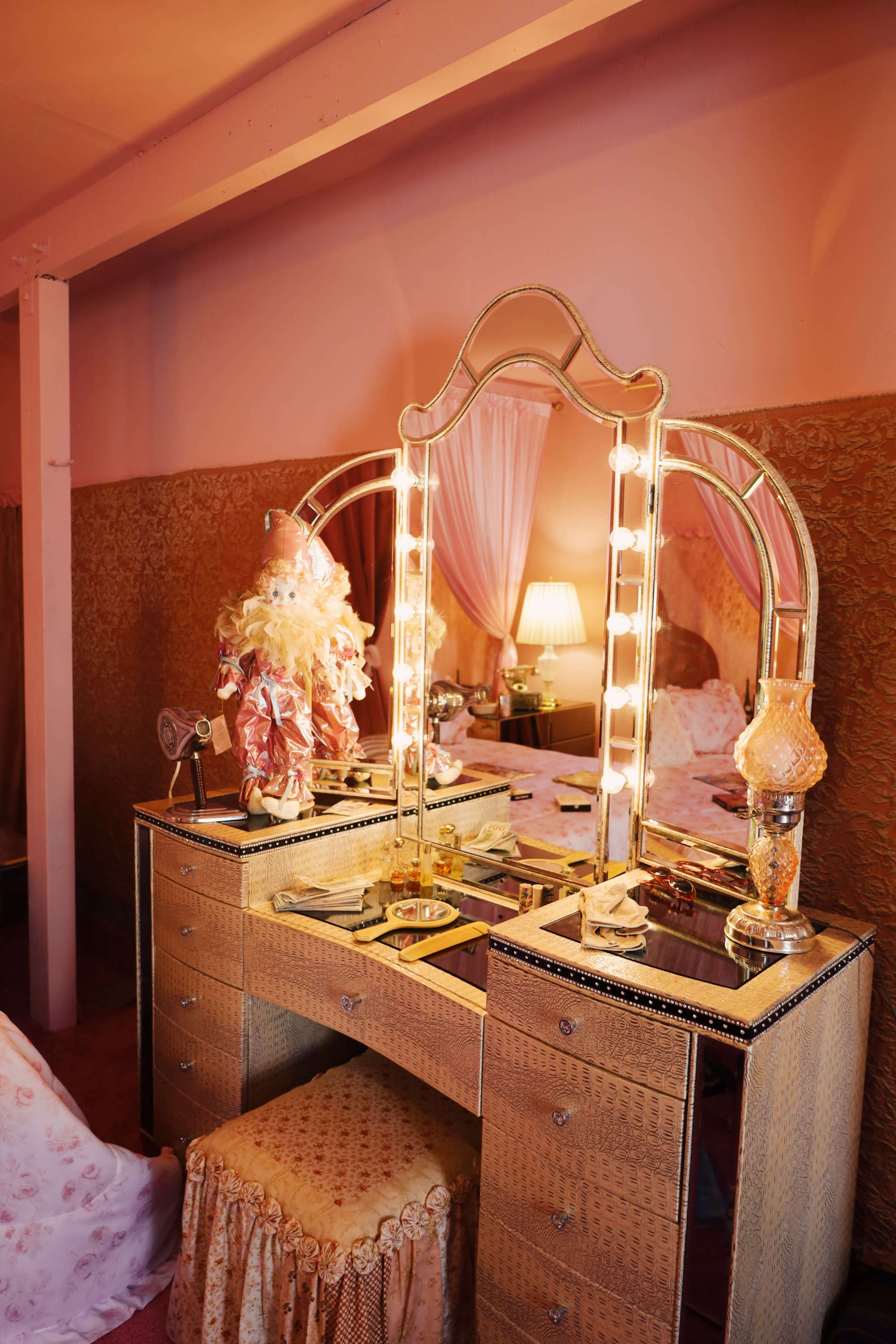 A vintage vanity table with a large mirror, surrounded by lamps and decorative items, set in a softly lit room.