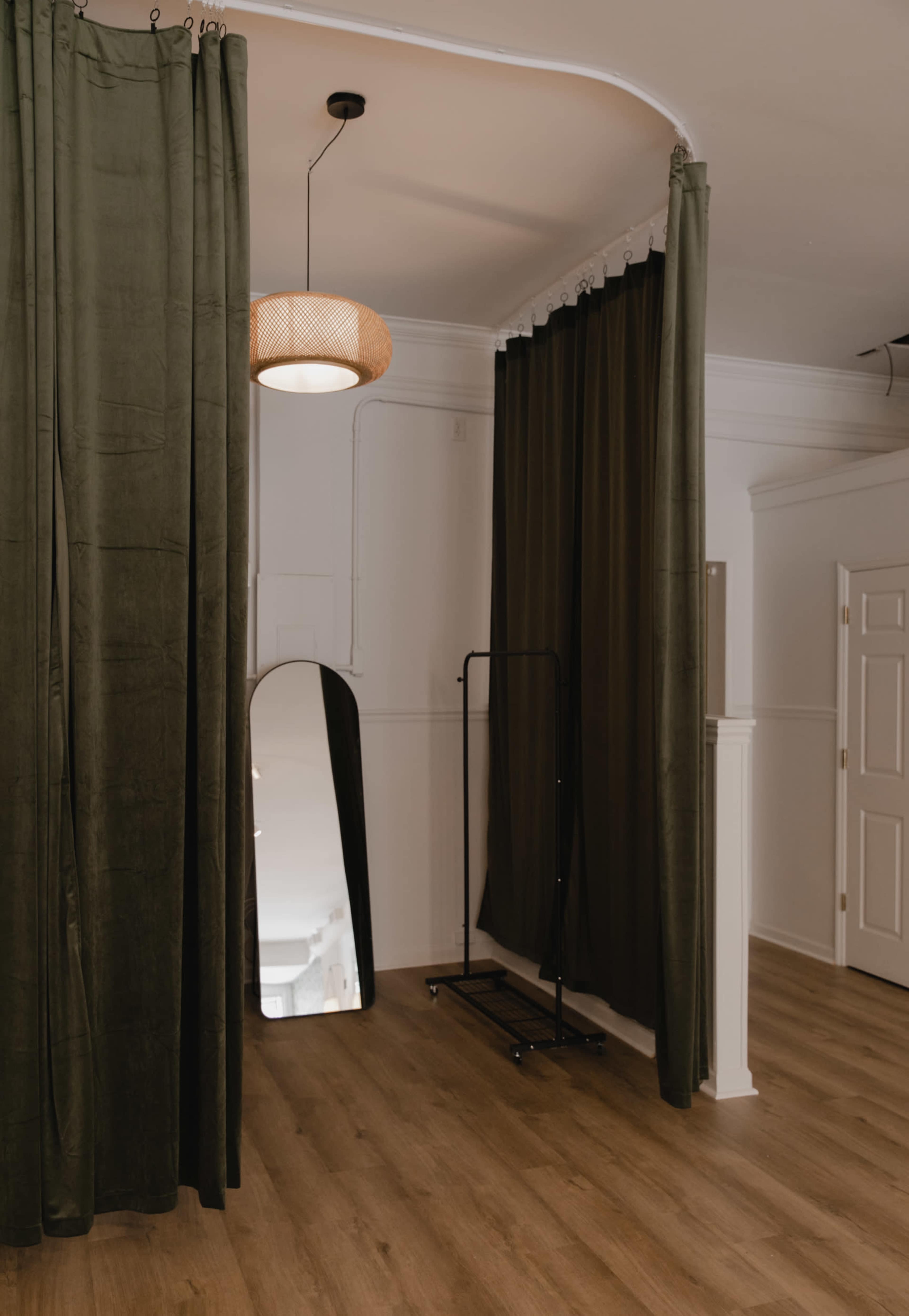 A room with green curtains, a mirror, a clothing rack, and a light fixture hanging from the ceiling.