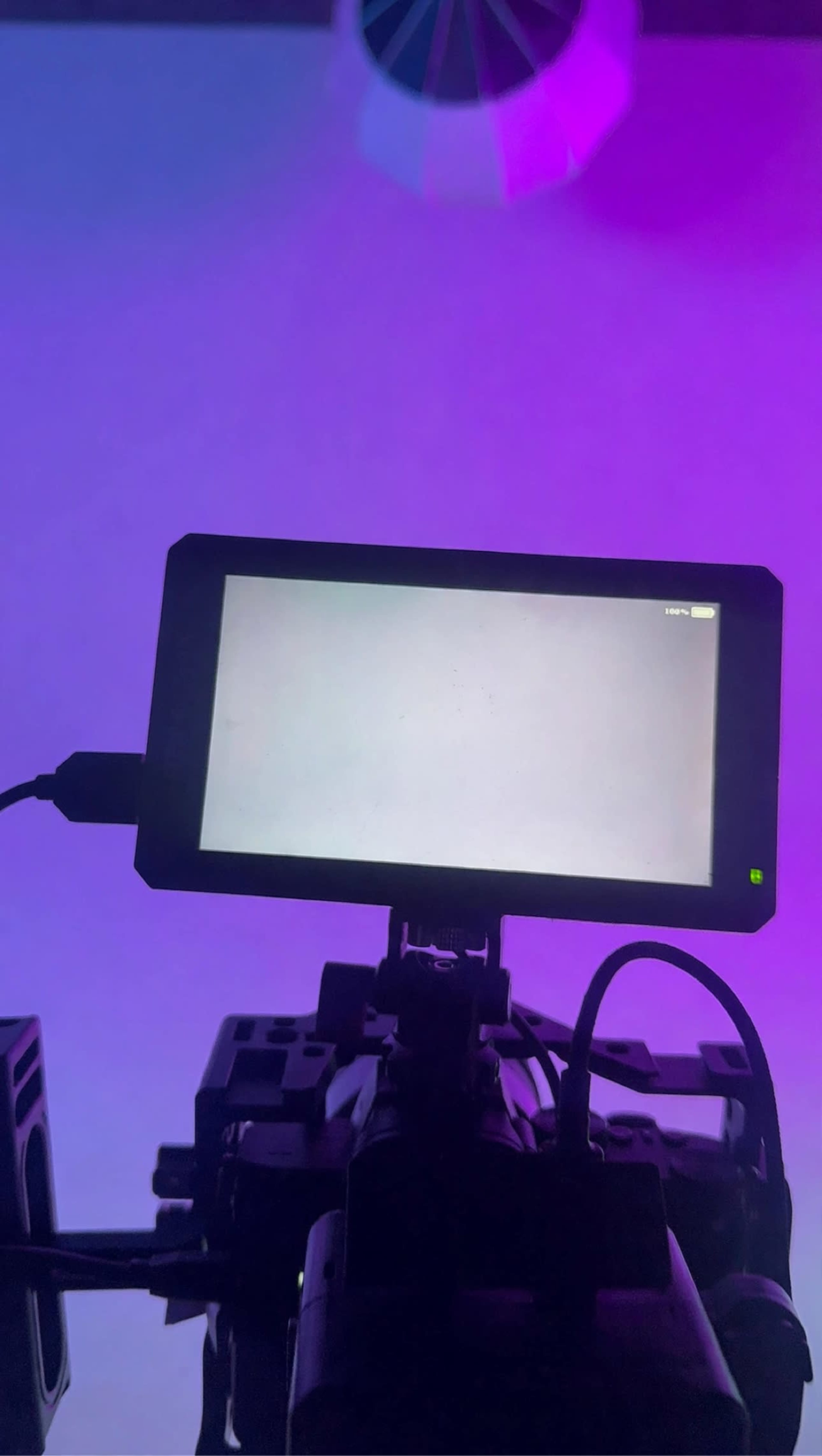 A camera monitor displays a blank screen, connected to a camera setup with purple lighting in the background.