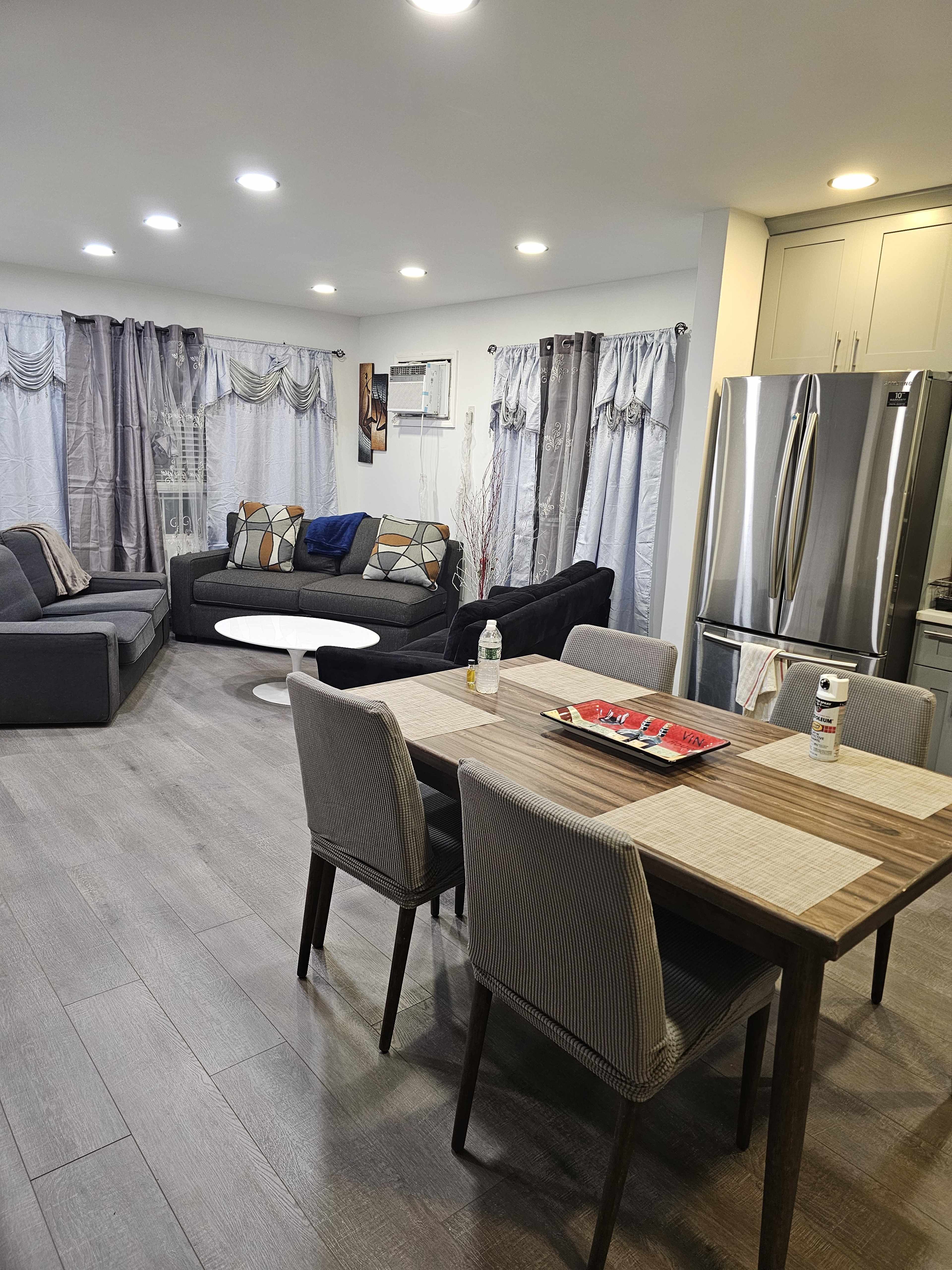 A modern living room and dining area with a gray sofa, a round coffee table, a dining table with chairs, and a stainless steel refrigerator in the kitchen area.